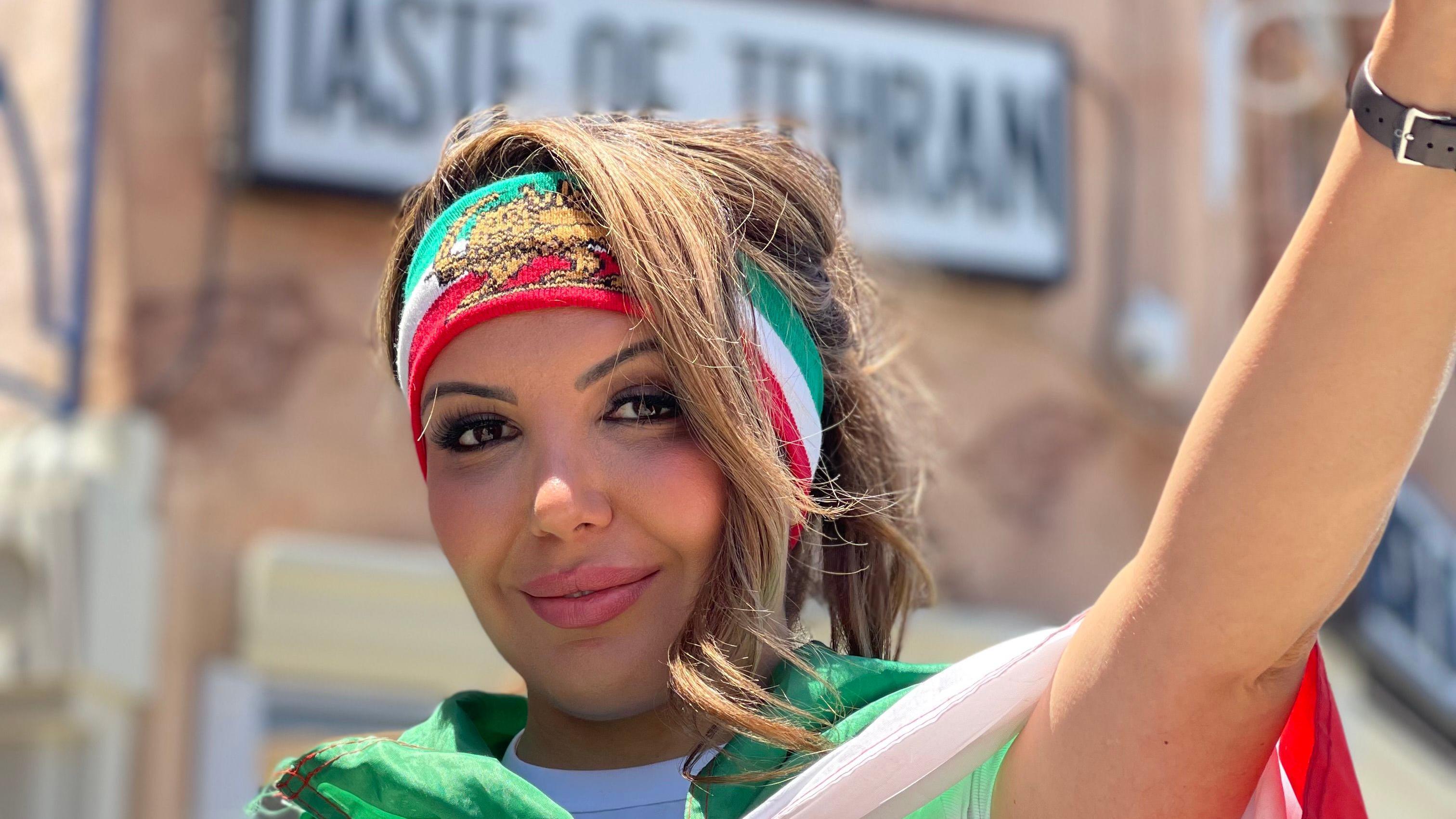 La iraní-estadounidense Paris Hobbs posa haciendo el gesto de la victoria con su mano izquierda, con la bandera previa a la Revolución Islámica de Irán con el león y el sol en el centro, que lice también en una cinta en la frente, afuera del restaurante Taste of Tehran en Los Ángeles, California, Estados Unidos, el 15 de junio de 2026. (Foto: Leire Ventas para BBC News Mundo).
