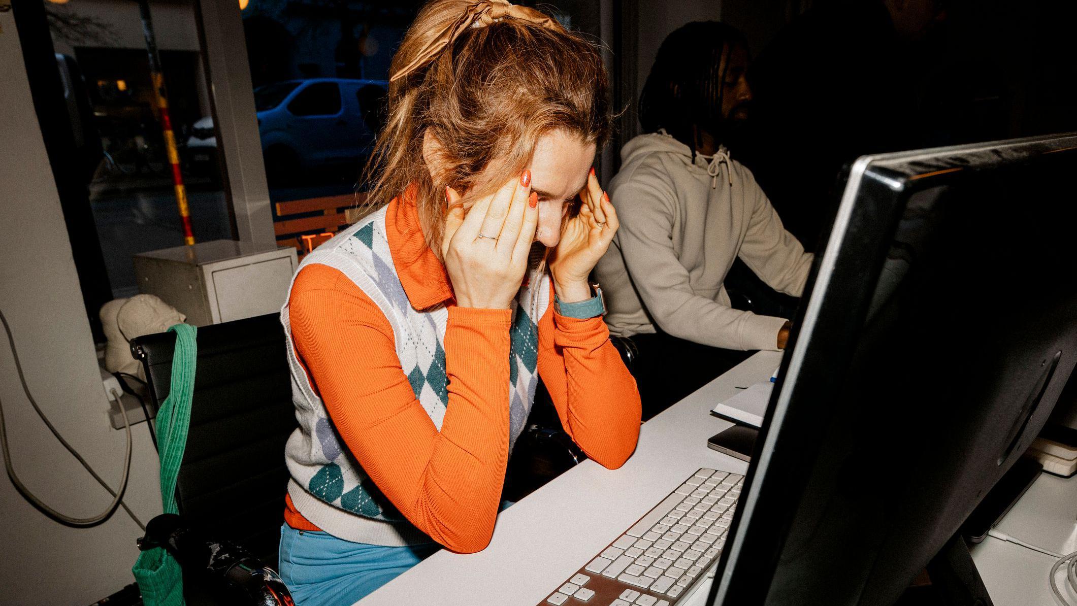 امرأة جالسة على كرسي ترتدي قميصاً برتقالياً بأكمام طويلة وسترة على مكتب عملها وتضع كلتا يديها على رأسها. 