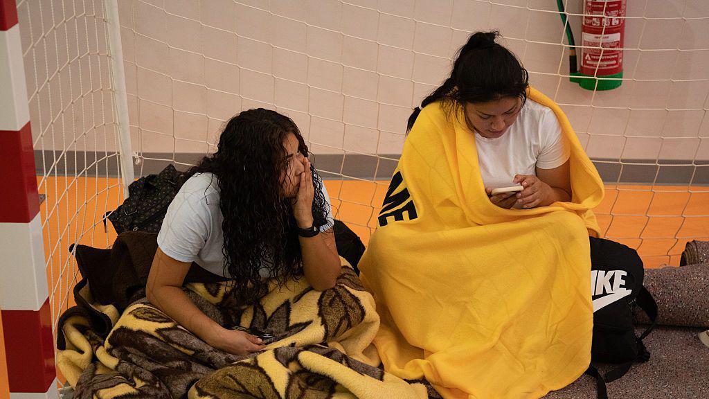 Dos mujeres dentro de una portería de fútbol sala, en manga corta pero abrigafas en las piernas con mantas. Alrededor tienen algunos enseres en mochilas. 