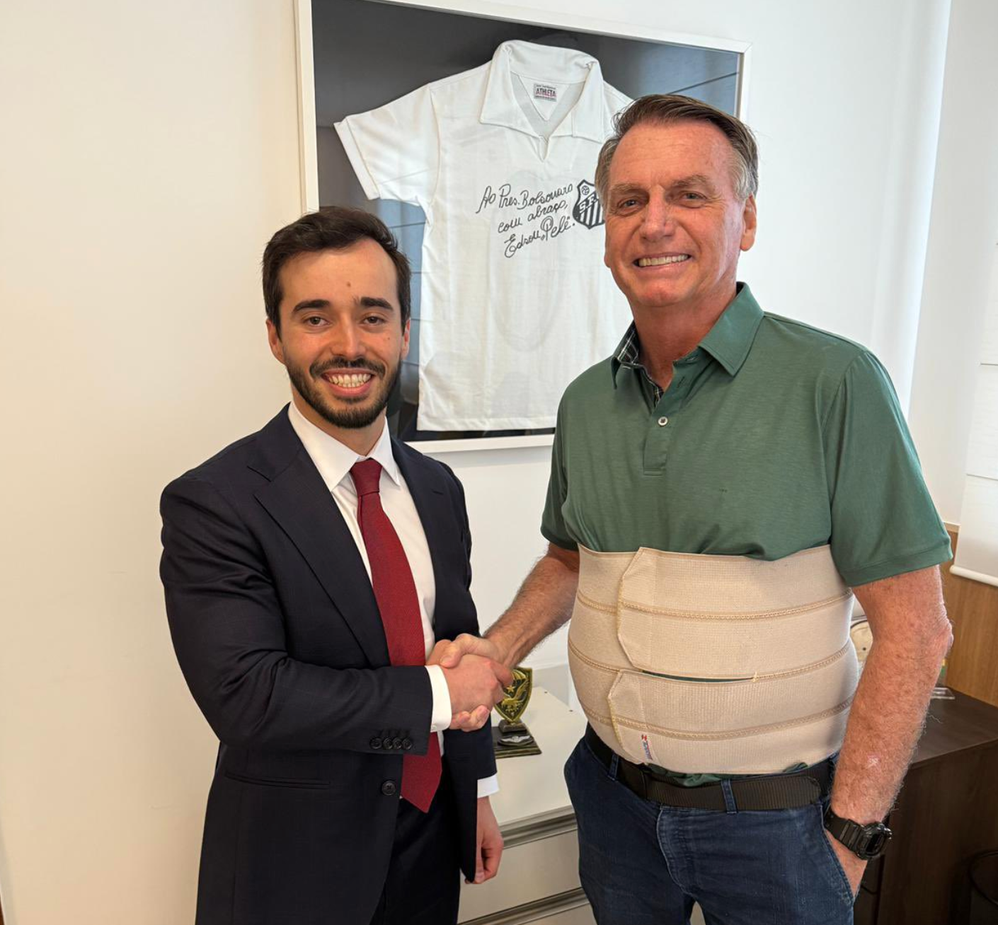 Foto de Ricardo Pita, usando terno azul marinho e gravata vermelha e Jair Bolsonaro, usando cinta de compress&atilde;o creme e camisa polo verde