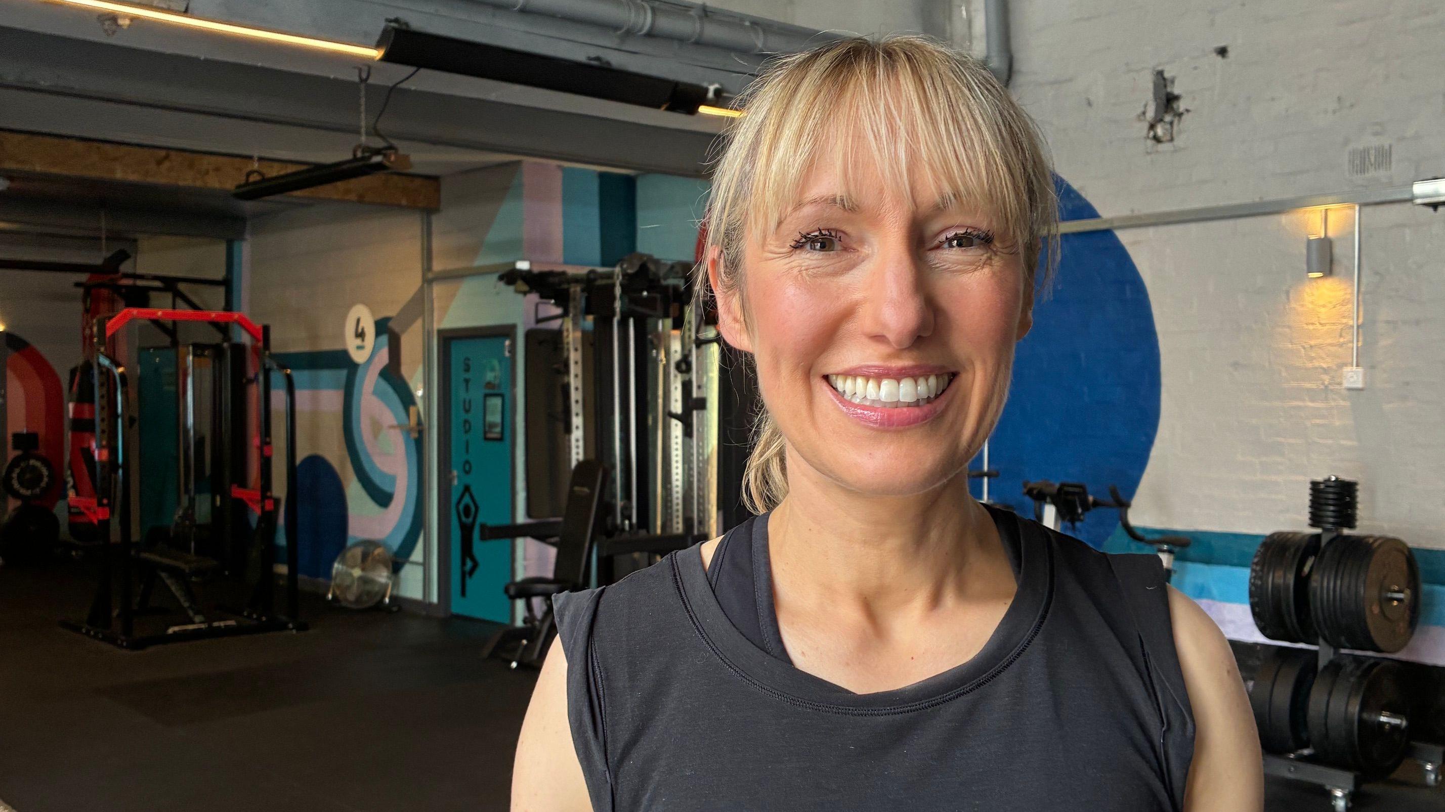 Bev Wilson est&aacute; em uma academia, vestindo roupas de gin&aacute;stica. Ela est&aacute; sorrindo para a c&acirc;mera e tem cabelos loiros em um rabo de cavalo.  