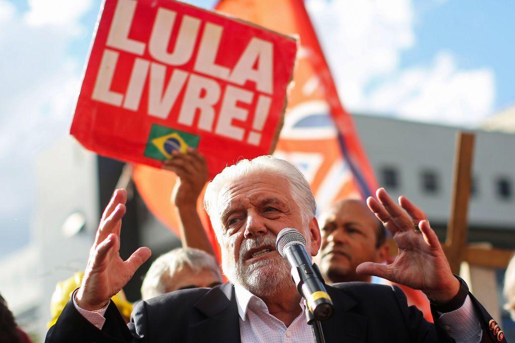 A imagem mostra Jaques Wagner discursando em um ato p&uacute;blico ao ar livre, falando ao microfone enquanto gesticula com as duas m&atilde;os erguidas. Ao fundo, apoiadores seguram bandeiras e um cartaz vermelho com a inscri&ccedil;&atilde;o &ldquo;LULA LIVRE!&rdquo;, al&eacute;m de uma pequena bandeira do Brasil.