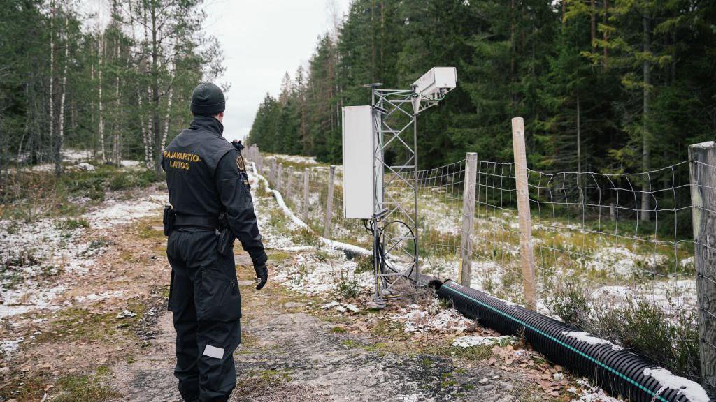 Militar uniformizado observa ao longo de uma cerca com postes de madeira, que divide um território coberto por um bosque com neve.