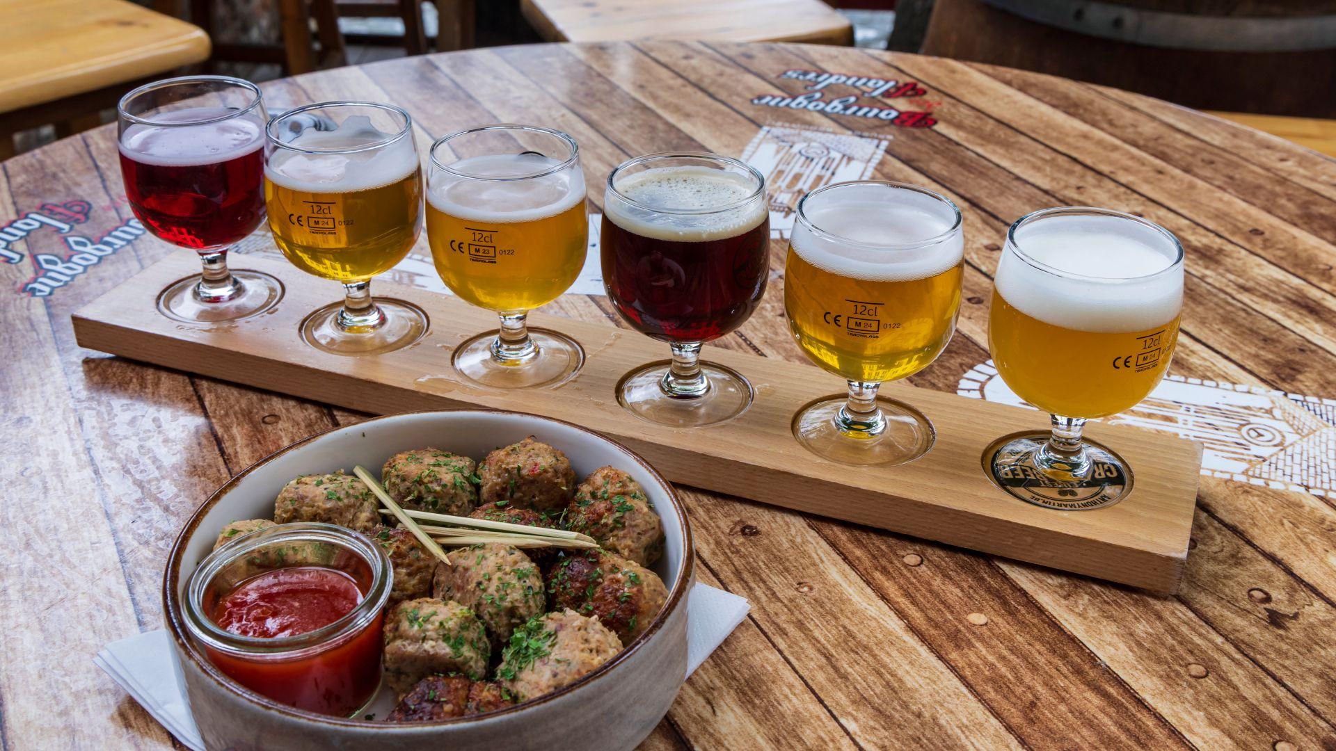 Drveni poslužavnik na kružnom drvenom stolu sadrži šest malih čaša piva, boje od crvene do zlatne, sa okruglim činijem sa ćuftama i sosom u prvom planu