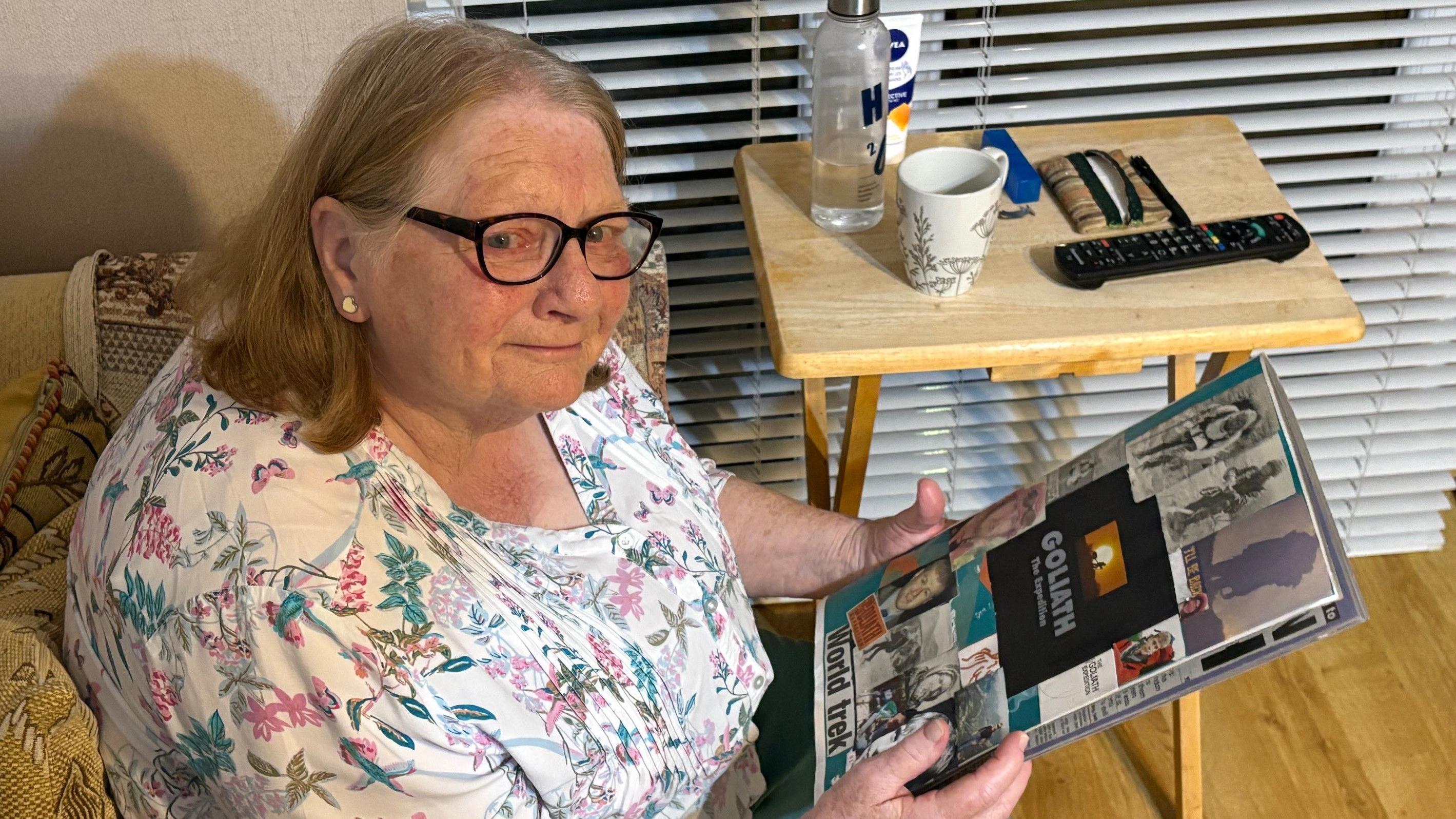 Uma mulher de &oacute;culos, com cabelos castanho-claros j&aacute; grisalhos, segura um &aacute;lbum de recortes com fotos do filho coladas na capa