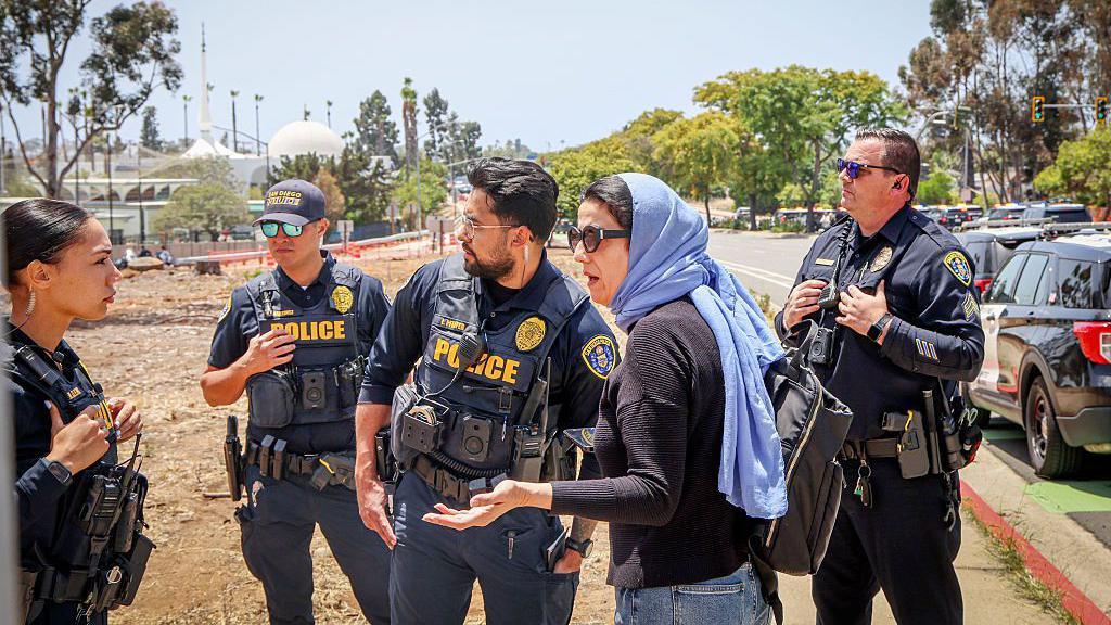 Seorang perempuan dengan jilbab biru pucat dan sweter hitam berbicara kepada sekelompok empat polisi berseragam di sebuah jalan