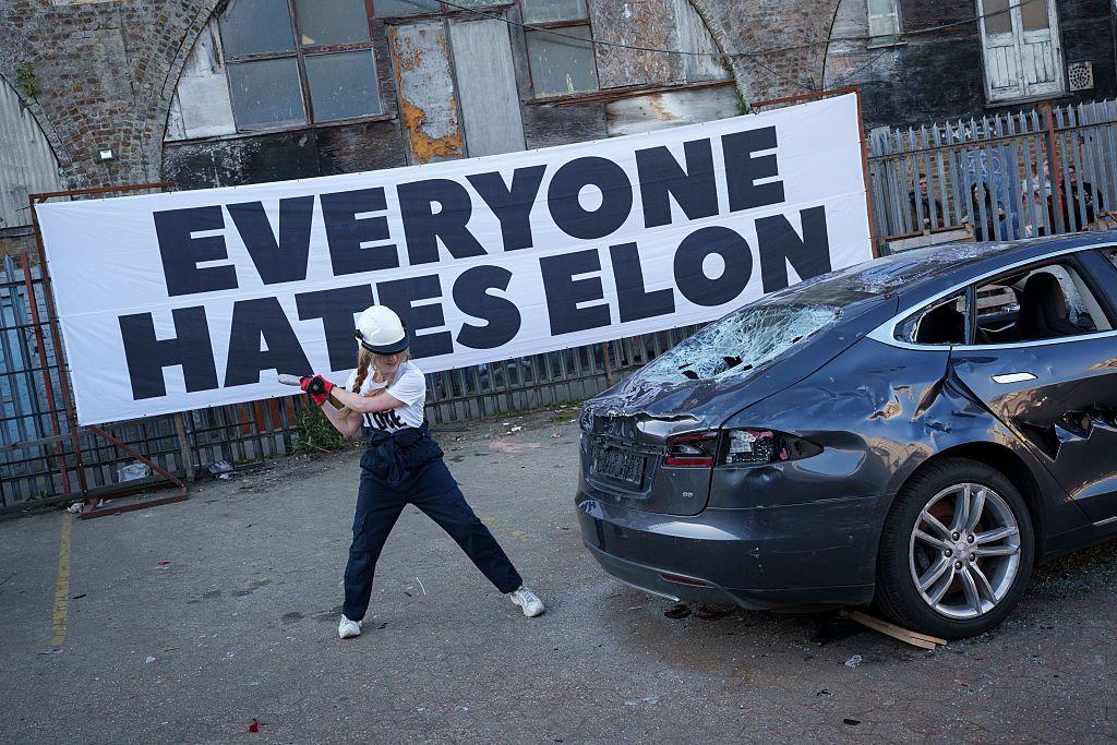 Una mujer golpeando un Tesla con un martillo, con un gran letrero atrás que dice "Todo el Mundo odia a Elon".