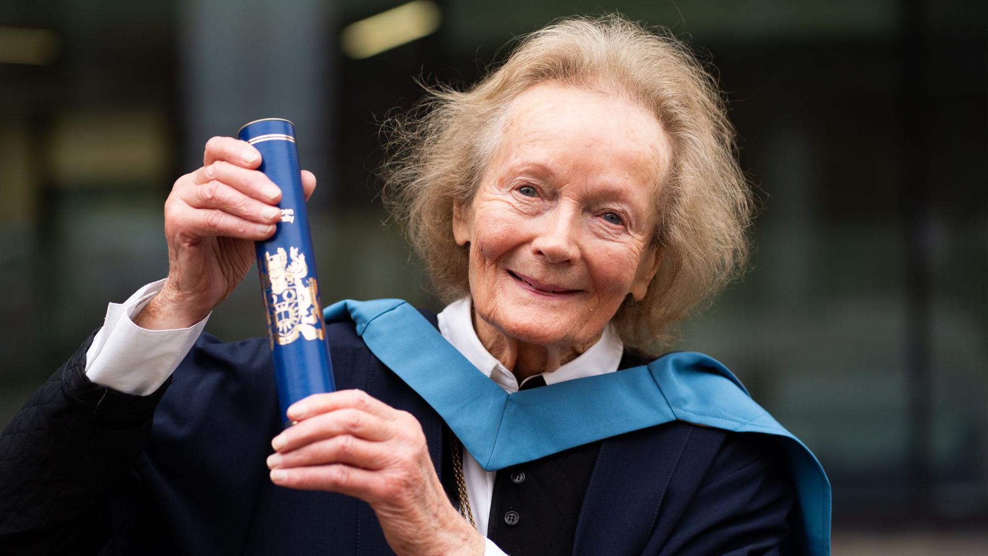 An older woman wearing graduation robes and holding a rolled up certificate. She has short grey hair.