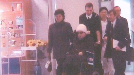 Ko Yong-hui, de &oacute;culos escuros e gorro branco, est&aacute; sentada em uma cadeira de rodas empurrada por um homem. V&aacute;rias pessoas est&atilde;o ao seu redor em um hospital na Fran&ccedil;a.