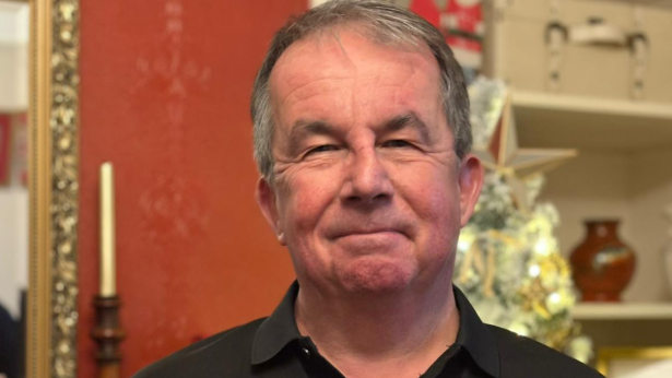 A man in his 60s with grey hair, wearing a black polo shirt. He is stood in front of a red wall, with a gold-framed mirror, a Christmas tree and a white book shelf with ornaments on it also visible.