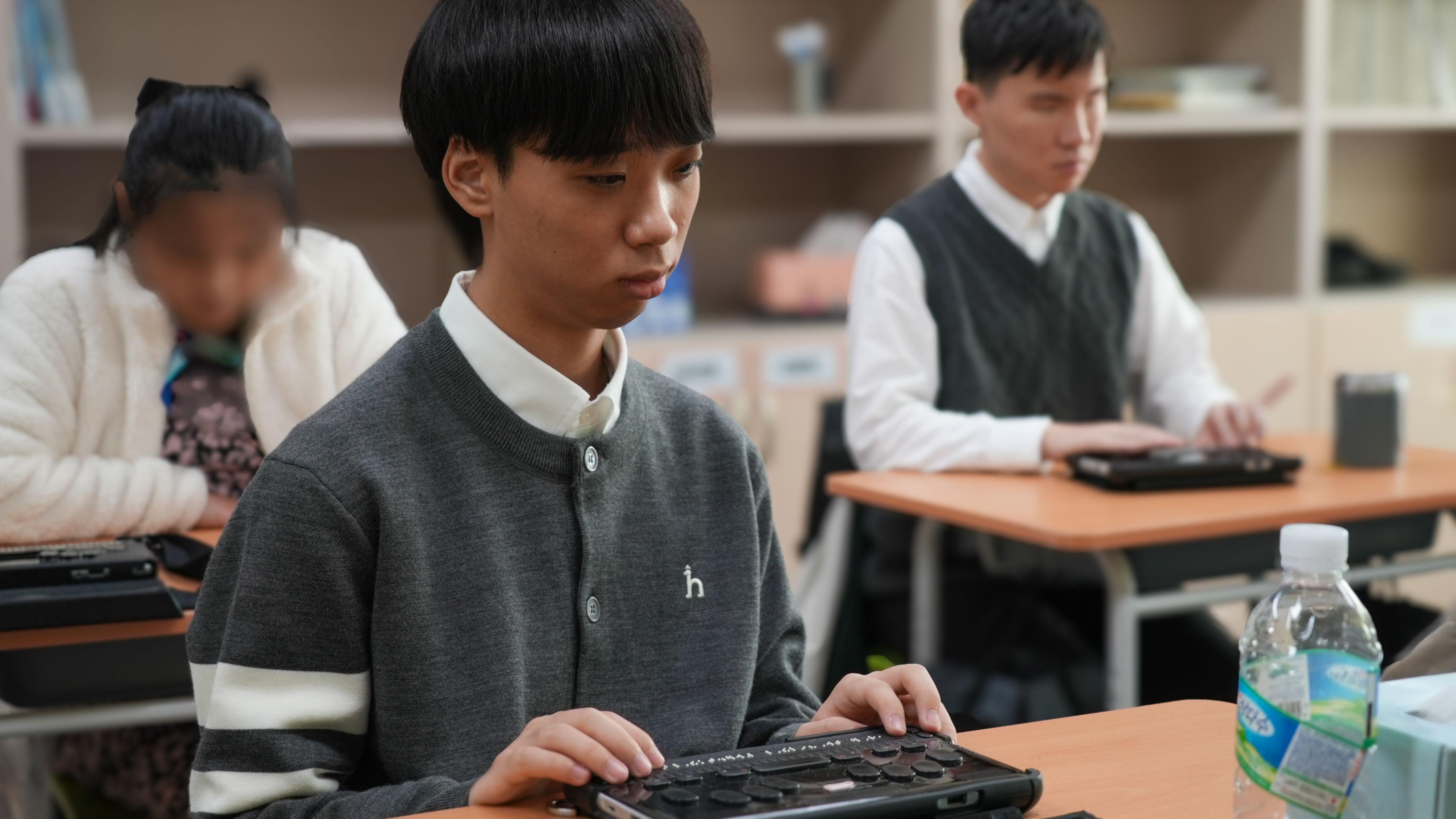 Students sit at their desks with their hands placed on braille display devices