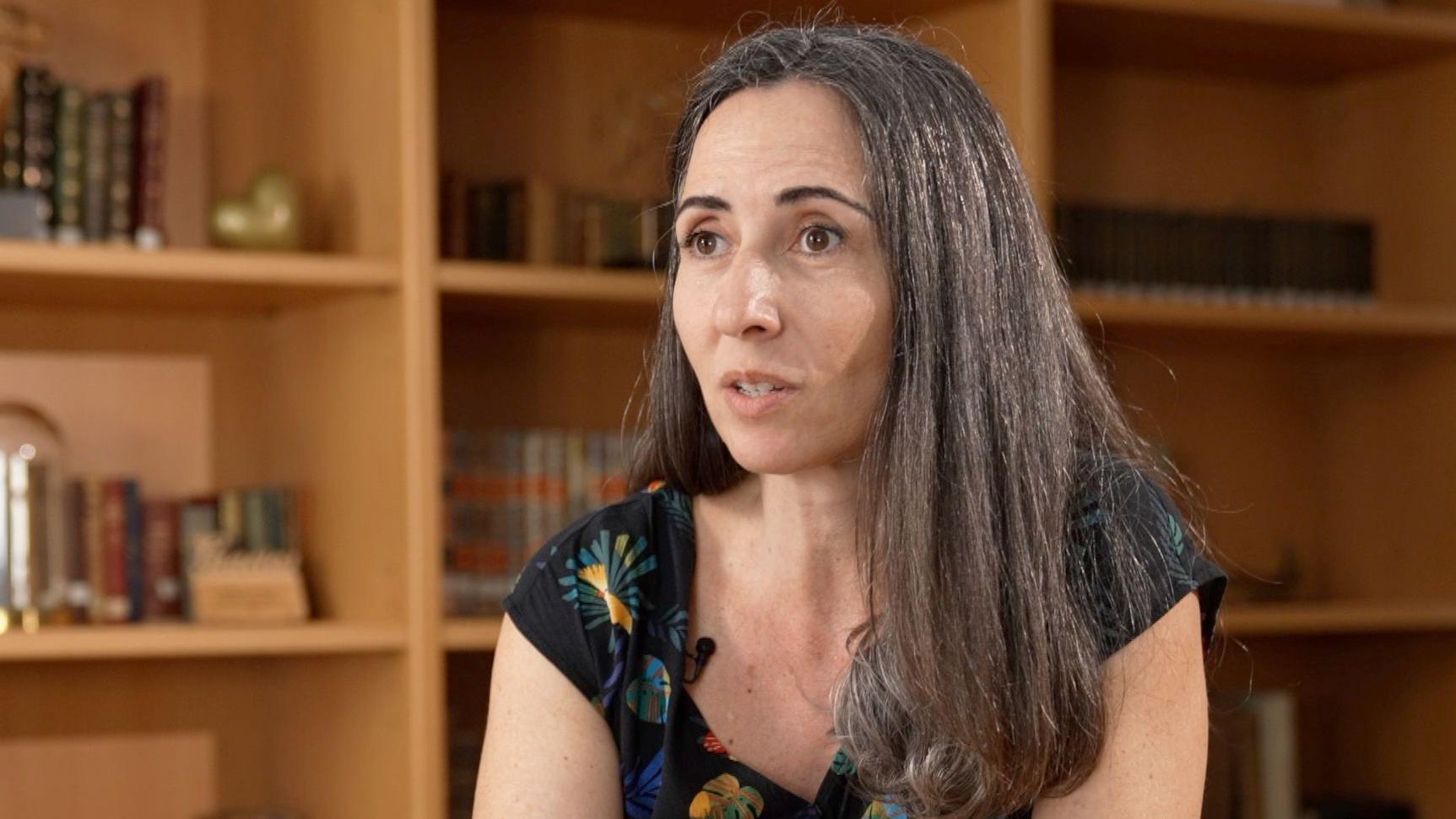 Carmona veste um vestido estampado e tem cabelos longos e castanhos; ela est&aacute; sentada perto de uma estante cheia de livros