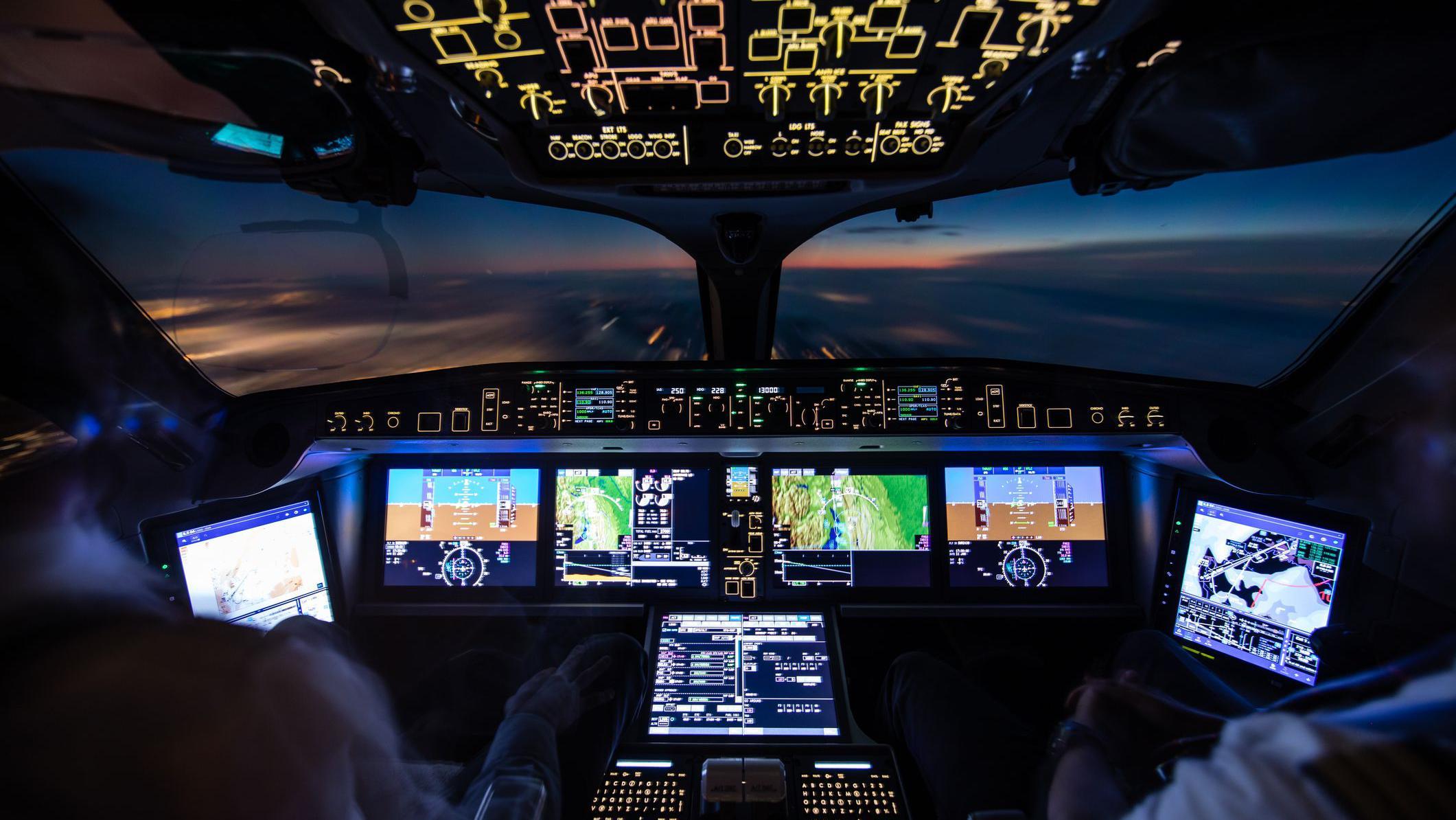 A imagem mostra o interior de uma cabine de comando moderna durante um voo ao amanhecer ou entardecer. Os pain&eacute;is de instrumentos e telas digitais iluminam o cockpit com tons de azul, verde e amarelo, exibindo informa&ccedil;&otilde;es de navega&ccedil;&atilde;o, altitude e sistemas da aeronave. Em primeiro plano, &eacute; poss&iacute;vel ver parcialmente dois tripulantes sentados diante dos controles. Pela ampla janela frontal, observa-se o horizonte com o c&eacute;u em tons azulados e alaranjados acima de uma camada de nuvens, criando uma atmosfera tecnol&oacute;gica e ao mesmo tempo contemplativa.