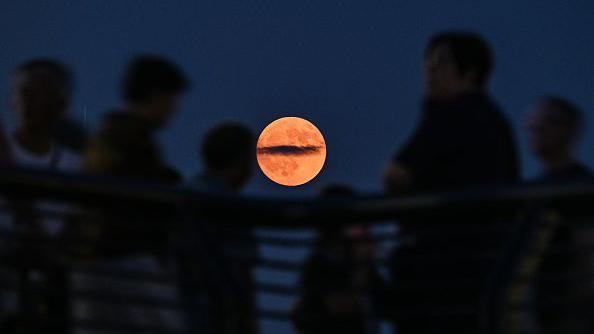 V&aacute;rias pessoas olhando para a Lua de Sangue, que tem uma cor laranja claro, no centro da foto.