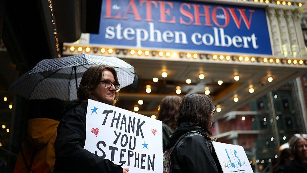 A imagem mostra um grupo de pessoas em frente ao teatro do programa &ldquo;The Late Show with Stephen Colbert&rdquo;, &agrave; noite e sob chuva. Em primeiro plano, uma mulher segura um guarda-chuva e um cartaz com a frase &ldquo;Thank You Stephen&rdquo;, ou &ldquo;obrigado, Stephen&rdquo;, em portugu&ecirc;s. Ao fundo, a marquise iluminada do programa aparece em destaque, cercada por luzes amarelas e pela movimenta&ccedil;&atilde;o da rua.