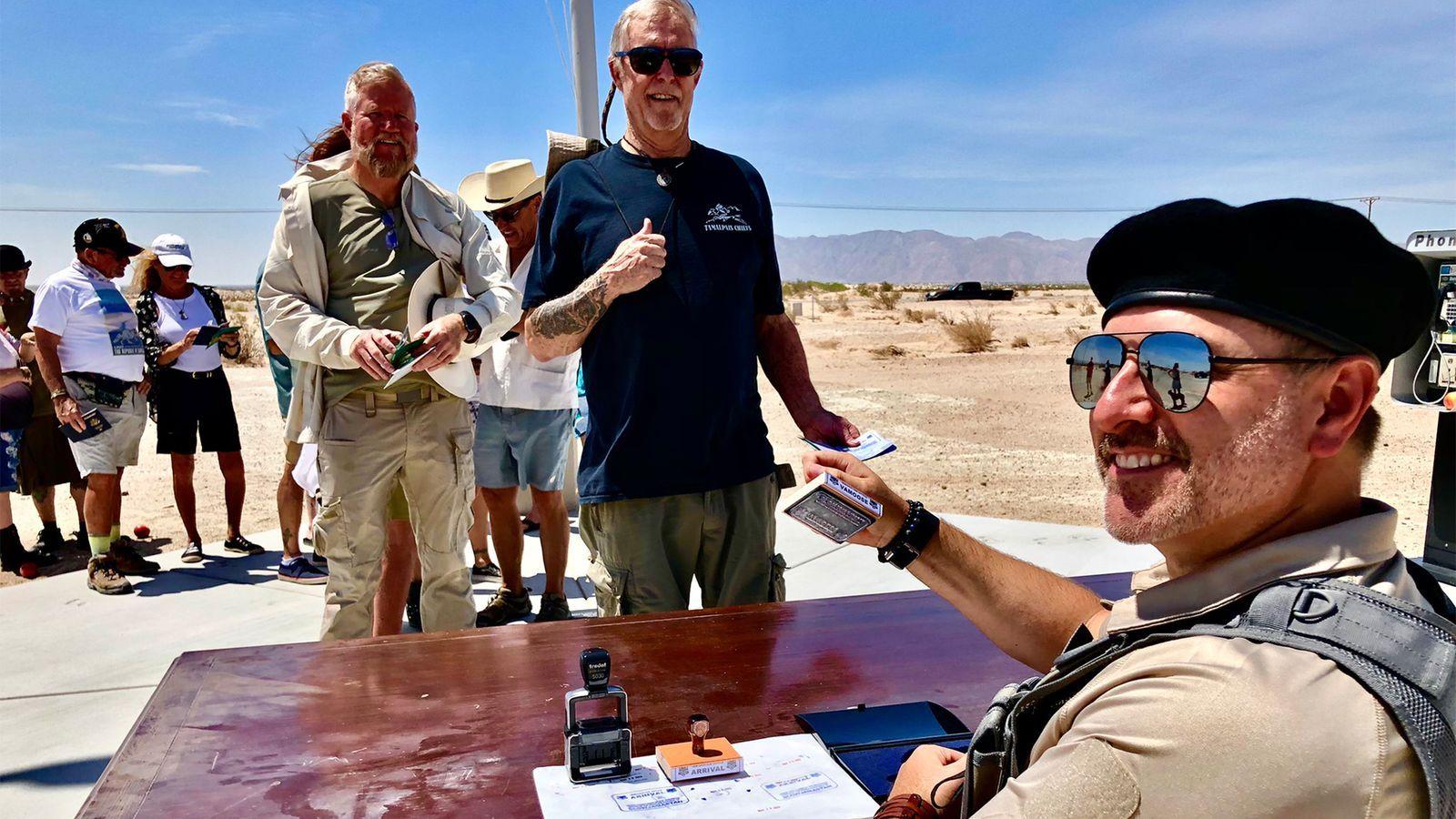 Un agente fronterizo con boina y gafas oscuras timbra los documentos de viaje de visitantes que espera en fila en el sol del desierto