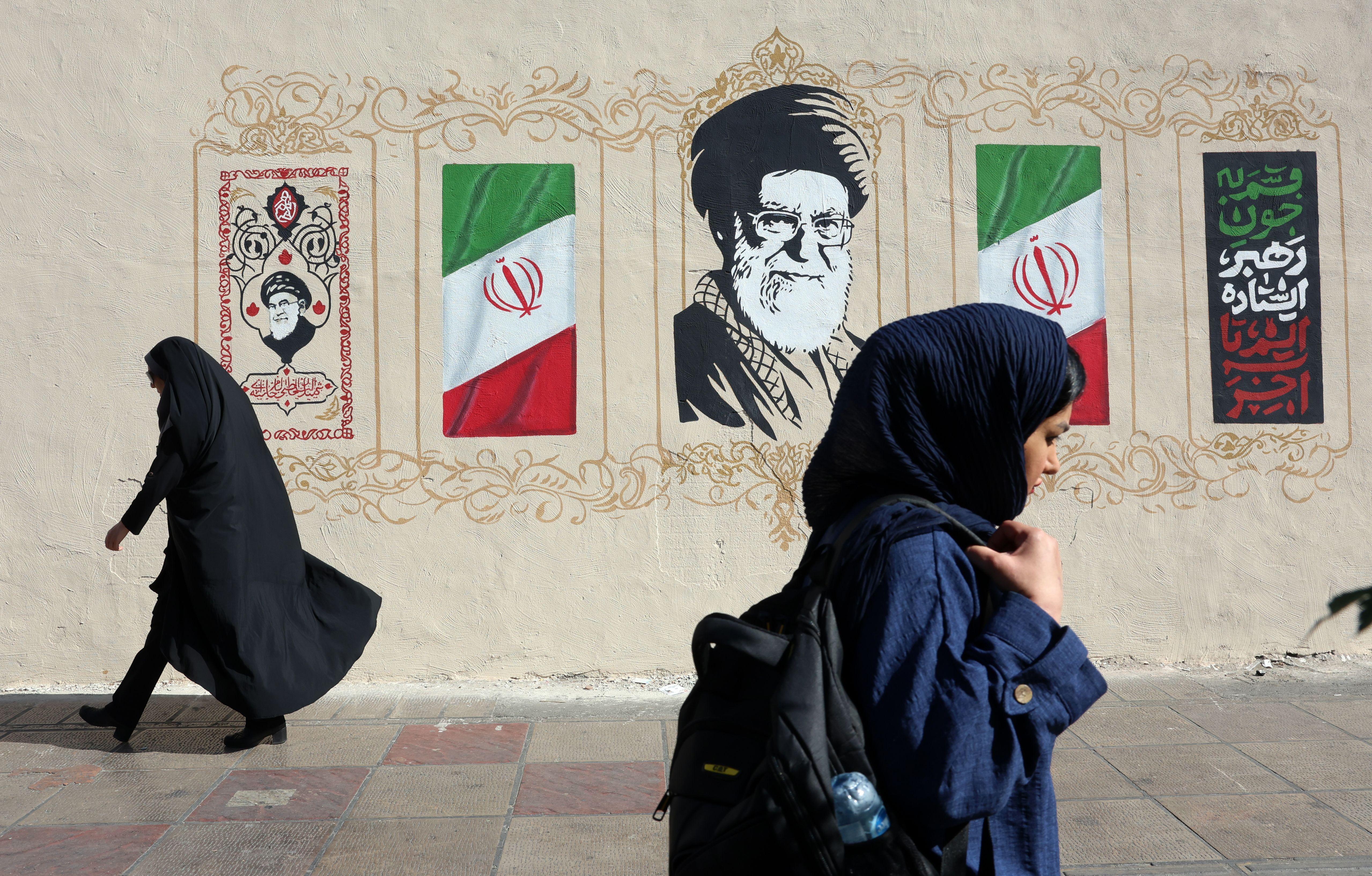 Dos mujeres con velo caminan por la acera frente a un mural que tiene banderas iraníes, caligrafía decorativa y un gran retrato de Alí Jamenei. Una mujer lleva una túnica negra, mientras que la otra viste un abrigo azul oscuro y lleva una mochila.
