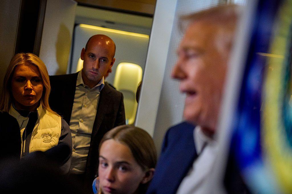 Trump, de perfil, habla con los periodistas en su avión presidencial, mientras una niña, su portavoz Karoline Leavitt y su asesor Stephen Miller lo escuchan desde un lateral.  