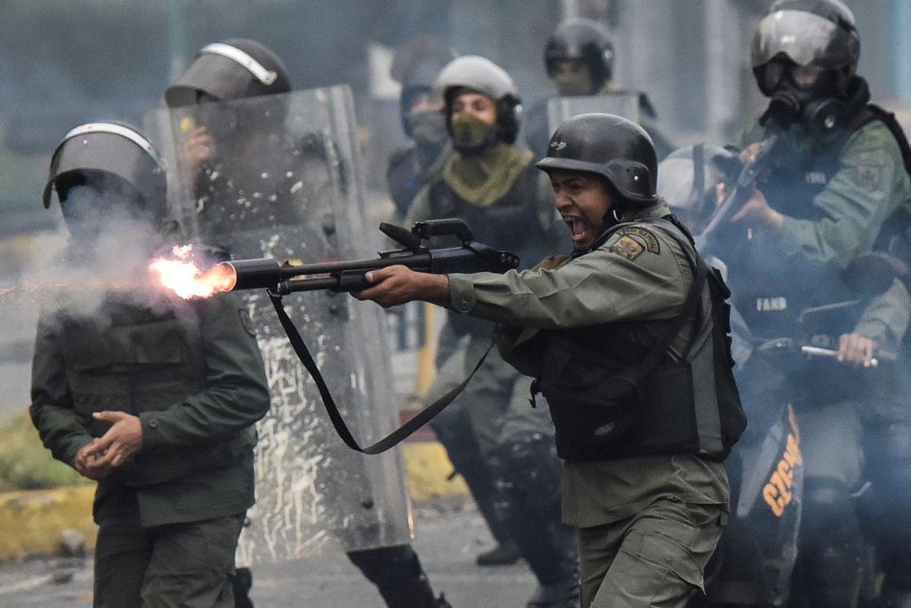 Un miembro de la Guardia Nacional dispara su escopeta contra manifestantes durante enfrentamientos. Está rodeado de otros miembros de la Guardia Nacional. Todos visten de color verde militar, con chaleco antibalas, casco. En muchos casos llevan la cara tapada. otros llevan un escudo transparente de protección. El militar que está en el centro sostiene un arma larga en sus manos. de la boca del arma se ve fuego. 
