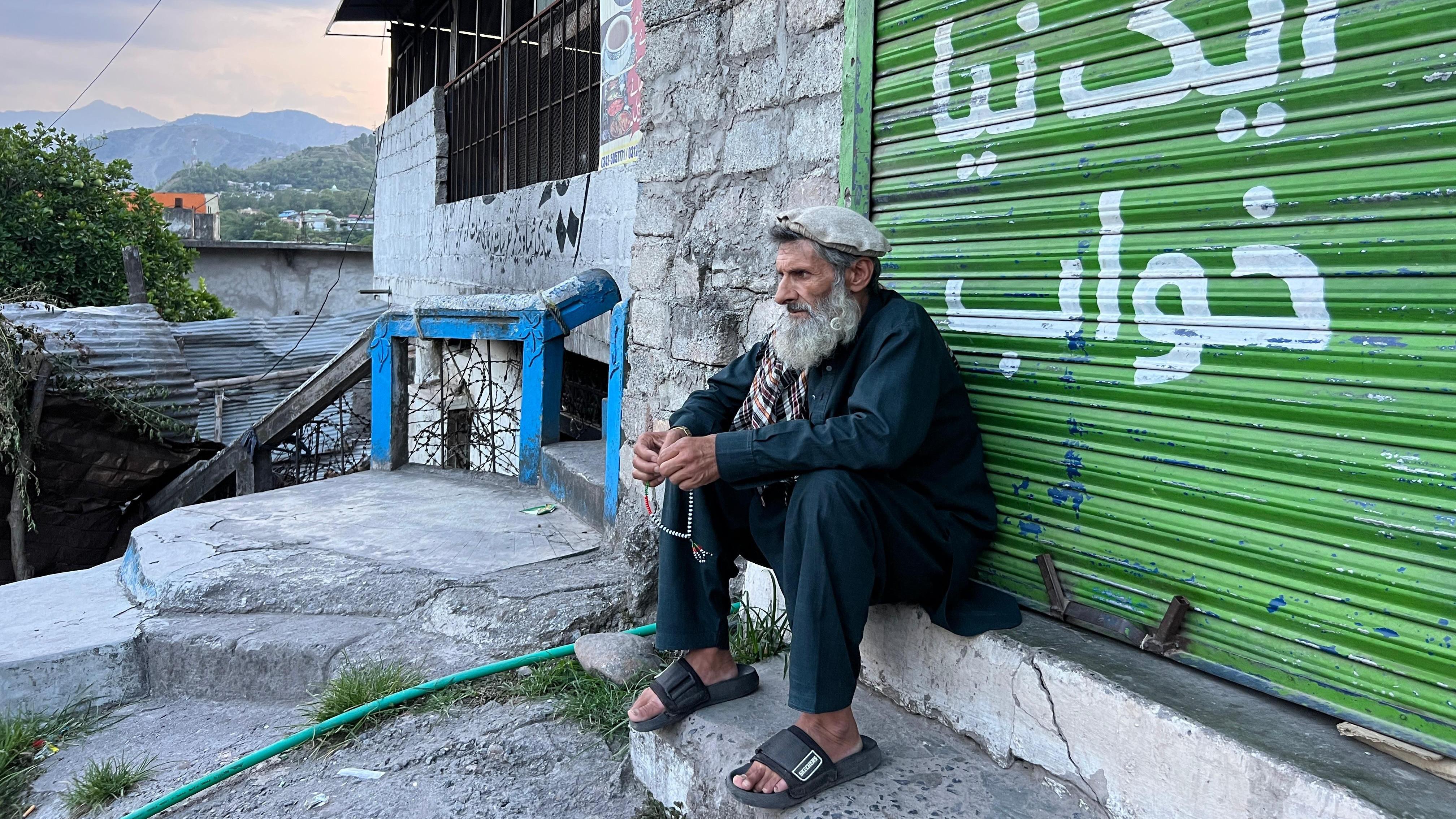 A man with a beard sits on a step in front of a bright green roller door. He is wearing a hat and flip-flops, and holding prayer beads in his fingers while staring forward pensively