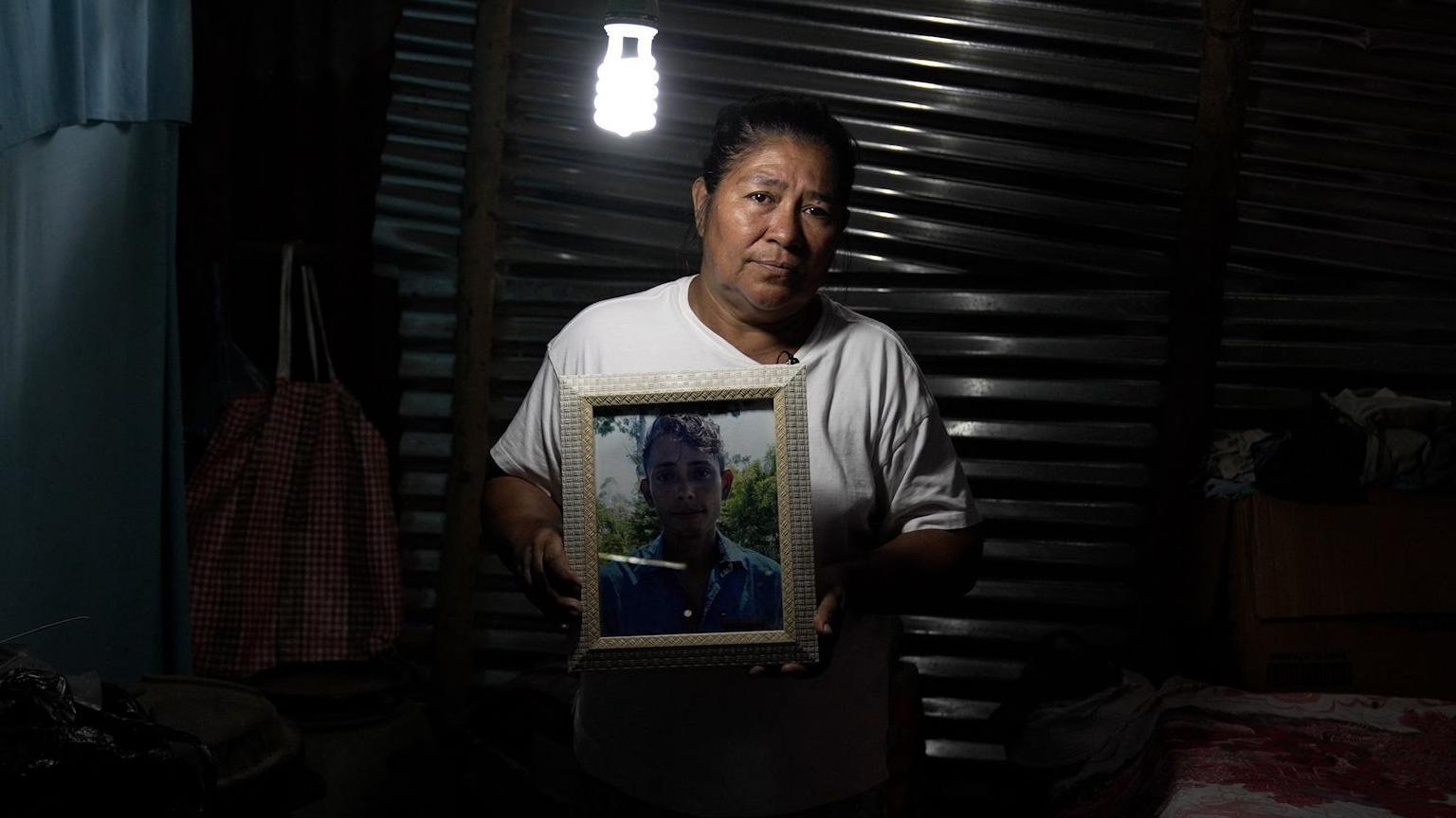 Una mujer sostiene el retrato de su hijo adulto. Está en un cuarto de una casa humilde, a media luz.