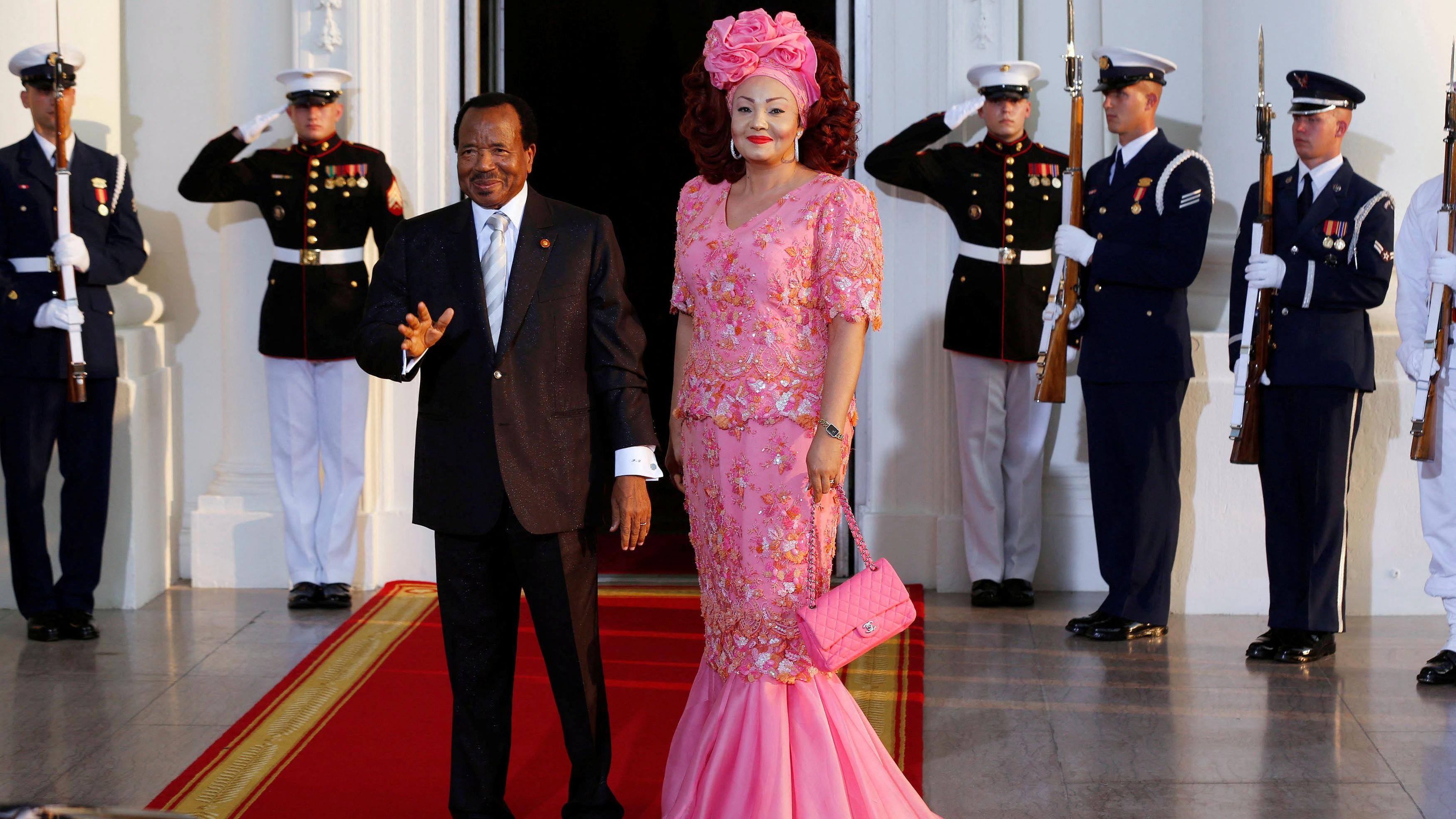 El presidente de Camerún con traje gris y corbata blanca, saluda a los fotógrafos. Su esposa, Chantal, con un vestido rosa floreado y un tocado haciendo juego, está junto a él.