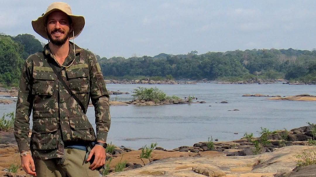 Rodrigo em pé, sorrindo, em frente a aparentemente uma lagoa ou rio