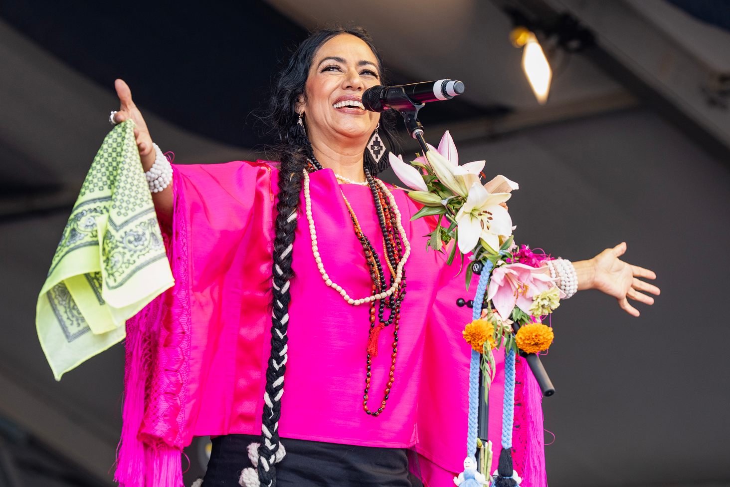 Lila Downs en el escenario frente a un micrófono. Lleva un poncho rosado, trenzas, collares de colores y en una mano tiene un pañuelo amarillo. Sonríe y tiene los brazos abiertos