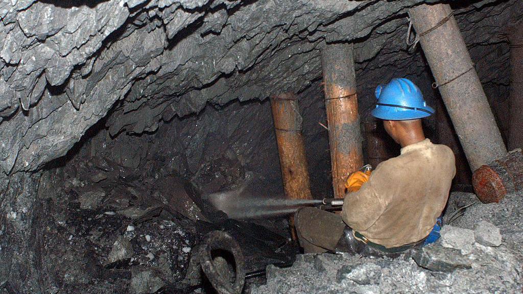 Un hombre con una camisa beige y un casco azul, de espaldas a la cámara, apunta un chorro de agua hacia una caverna rocosa dentro de una cueva.