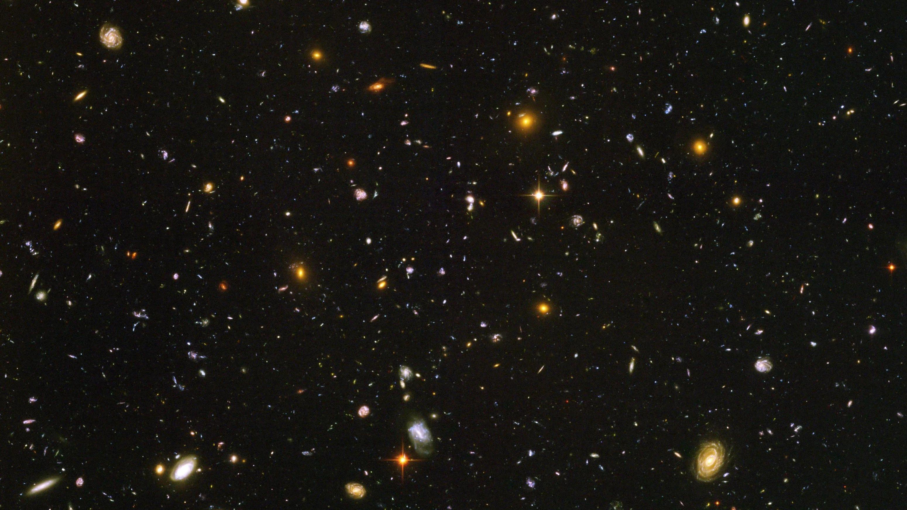 The image shows a completely black sky sprinkled with thousands of tiny coloured specks: every speck is a distant galaxy, not a single star.The galaxies come in many shapes and colours, from small bluish smudges to larger golden spirals and fuzzy red blobs, scattered evenly across the frame.There is no foreground or horizon, just a deep, seemingly endless view into the distant Universe packed with galaxies of all kinds.