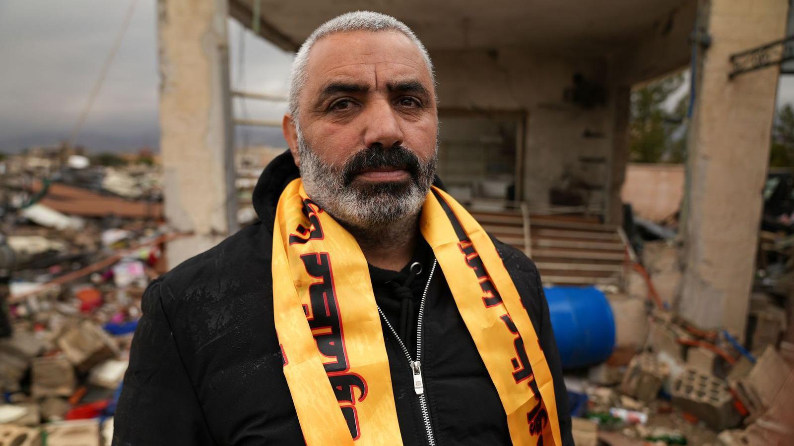 يقف حسن الطحان أمام المبنى المهدم وهو يرتدي وشاحاً أصفر اللون يحمل شعار حزب الله حول عنقه