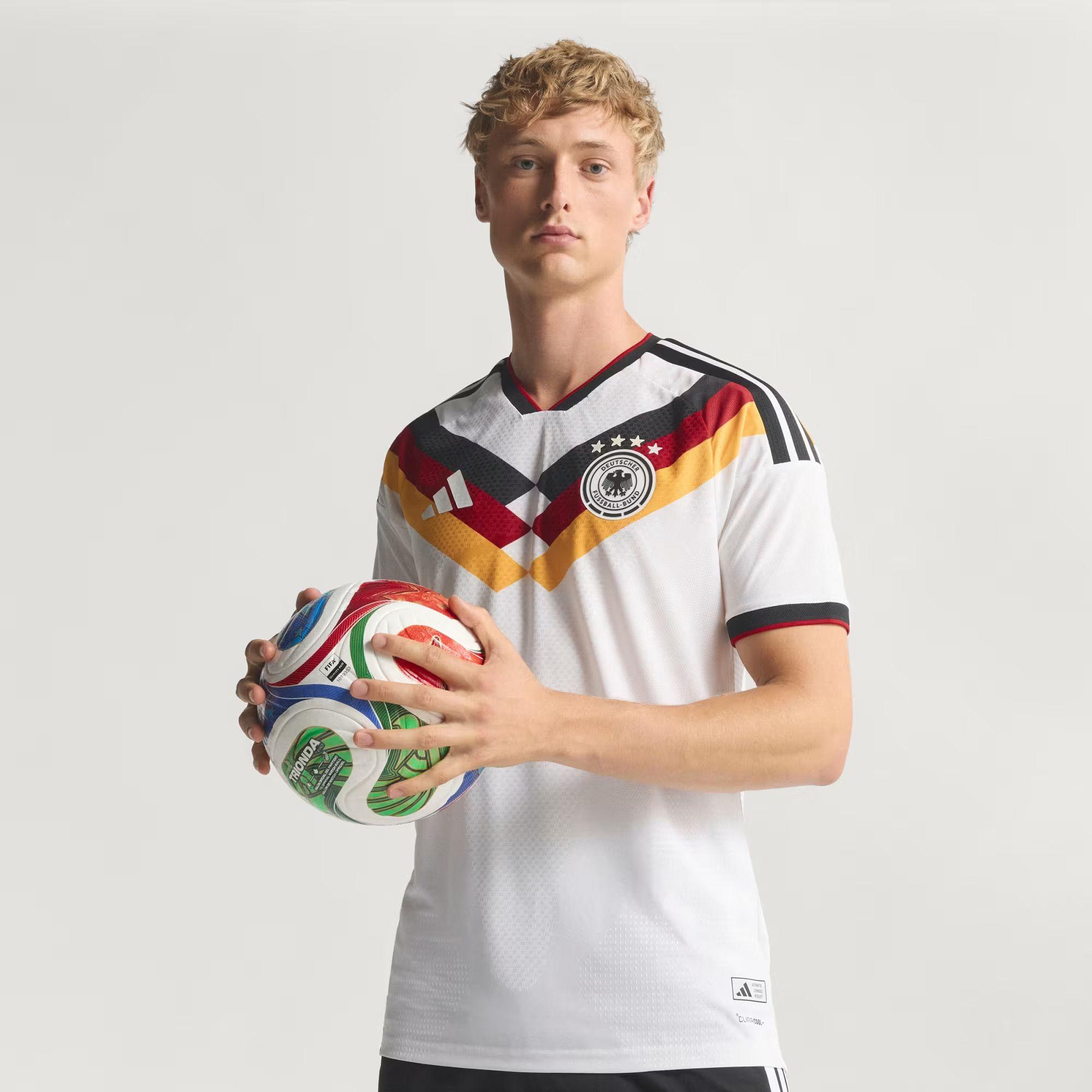 A imagem mostra um jovem segurando uma bola de futebol colorida com as duas m&atilde;os enquanto veste a camisa branca da sele&ccedil;&atilde;o da Alemanha. O uniforme tem detalhes geom&eacute;tricos em preto, vermelho e amarelo no peito, remetendo &agrave;s cores da bandeira alem&atilde;, al&eacute;m do escudo da federa&ccedil;&atilde;o do pa&iacute;s com quatro estrelas e o logotipo da Adidas.