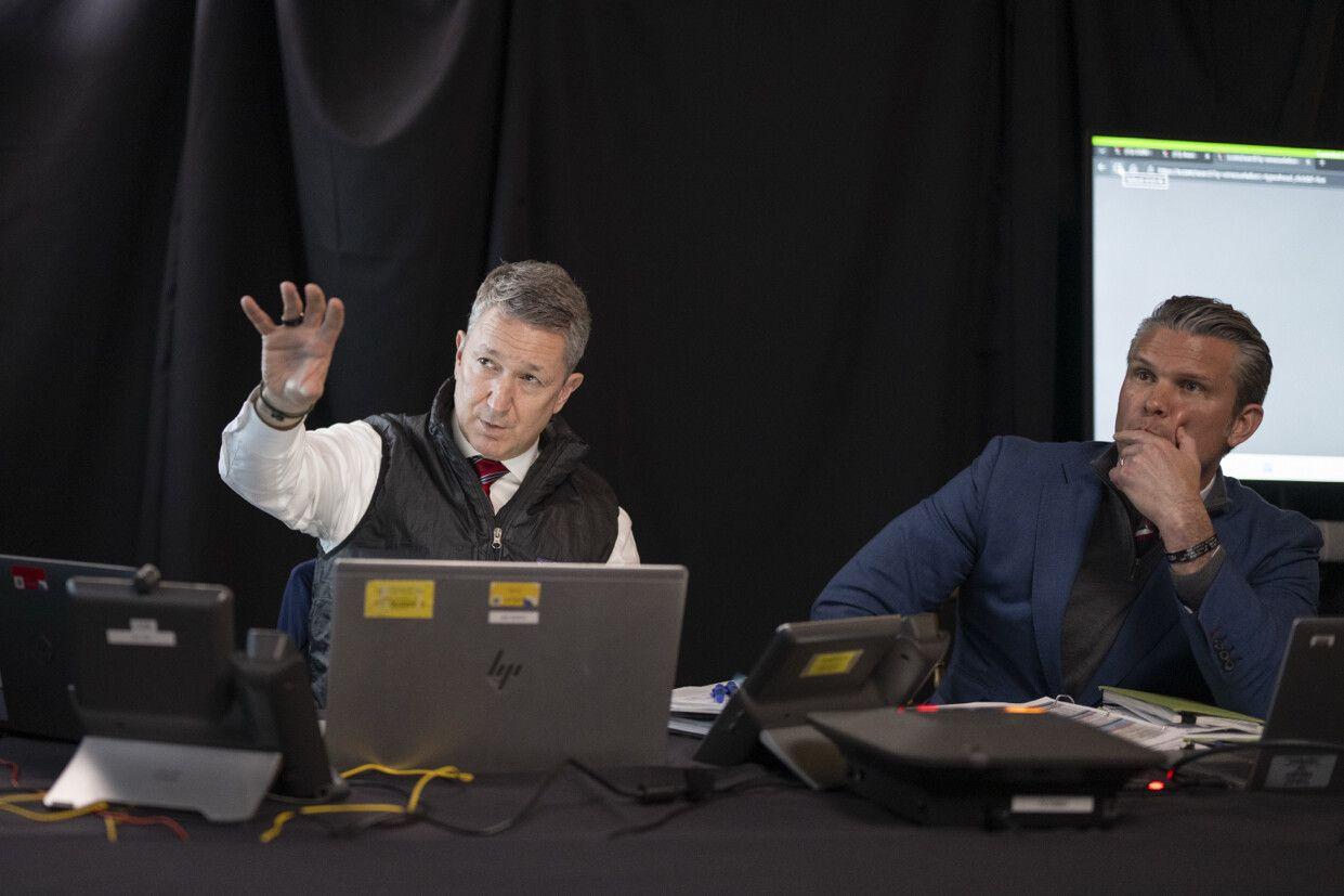 Un funcionario hace un gesto con la mano mientras mira hacia delante, y Hegseth mira atento con su mano izquierda sobre la parte inferior de su cara.