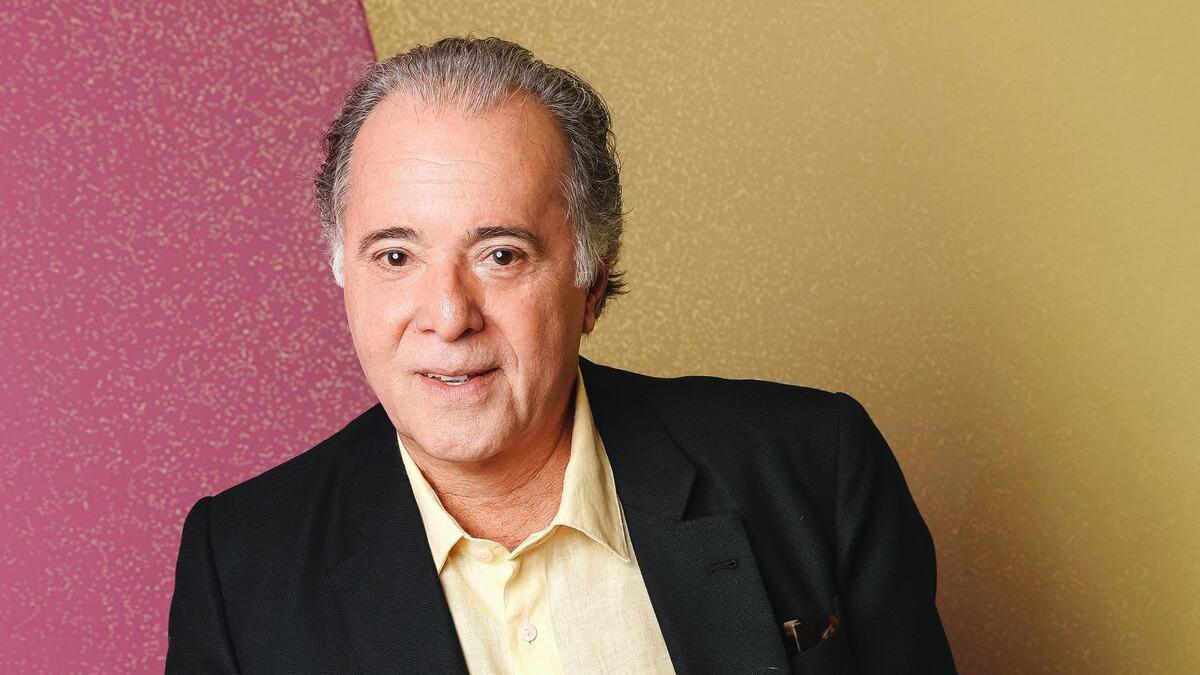 Tony Ramos, com cabelos grisalhos penteados para trás, aparece em um retrato posado. Ele veste blazer preto sobre uma camisa clara e olha diretamente para a câmera com expressão simpática. O fundo é dividido em duas cores — rosa à esquerda e amarelo à direita.