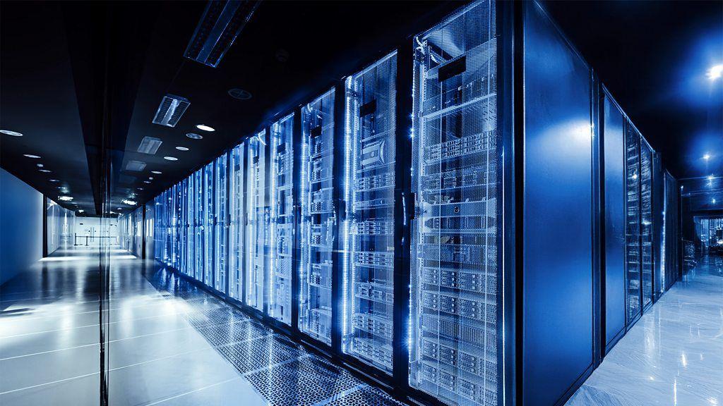 Imagen de un centro de datos. Un pasillo se bifurca hacia la izquierda y hacia la derecha. En el medio se ve una larga hilera de torres de color azules dentro de las cuales parece haber máquinas de procesamiento de datos.