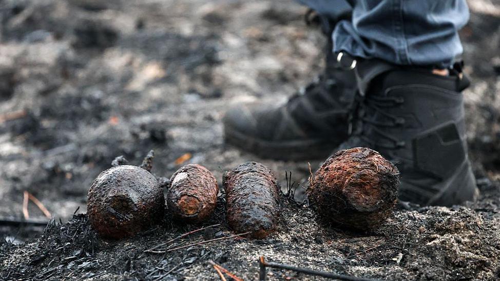 En primer plano, unas botas negras de oficial. A su lado hay cuatro objetos que parecen piñas pero que en realidad son proyectiles de la Segunda Guerra Mundial