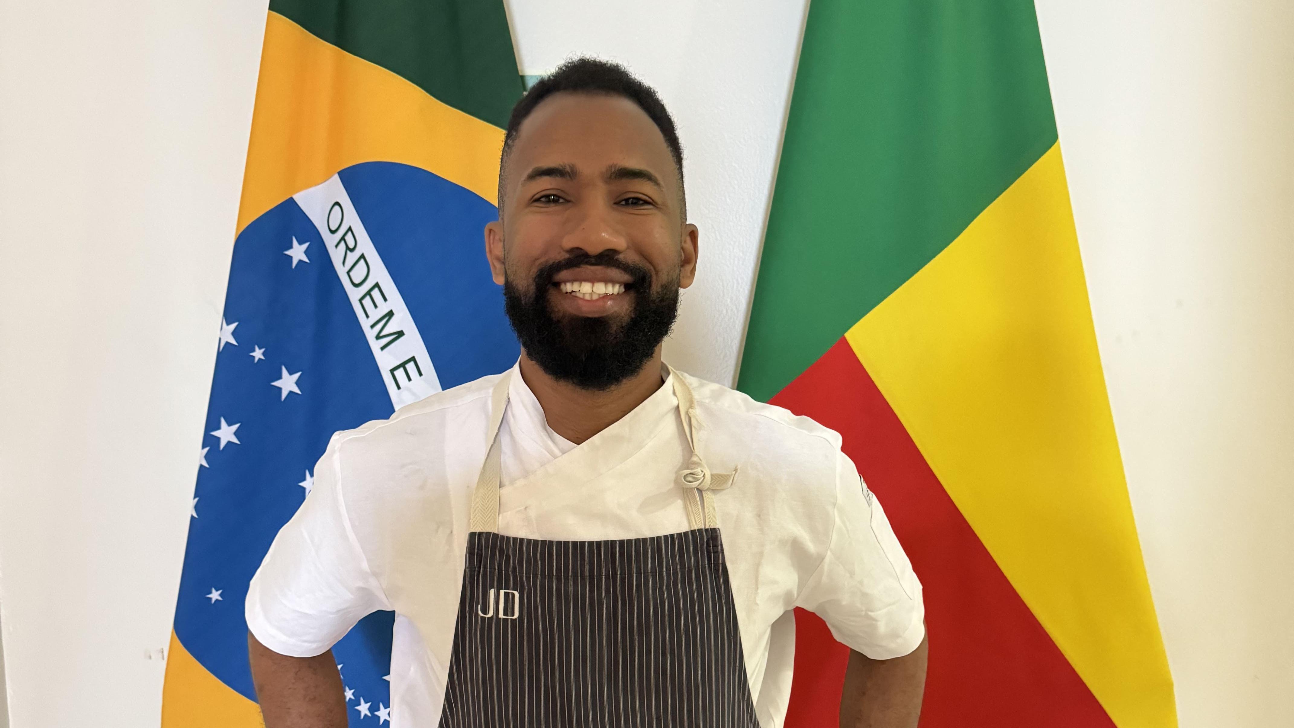 Homem com avental de cozinheiro sorrindo em frente a bandeiras do Brasil e Benin