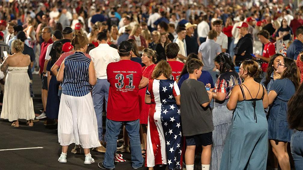 Un numeroso grupo de personas hace fila para entrar en el estadio State Farm en Glendale, Arizona, EE.UU. Una mujer tiene un vestido con la bandera de estrellas y franjas de Estados Unidos. Otro hombre de espaldas porta una camiseta roja con el número 31 que lee: "Gracias, Charlie".