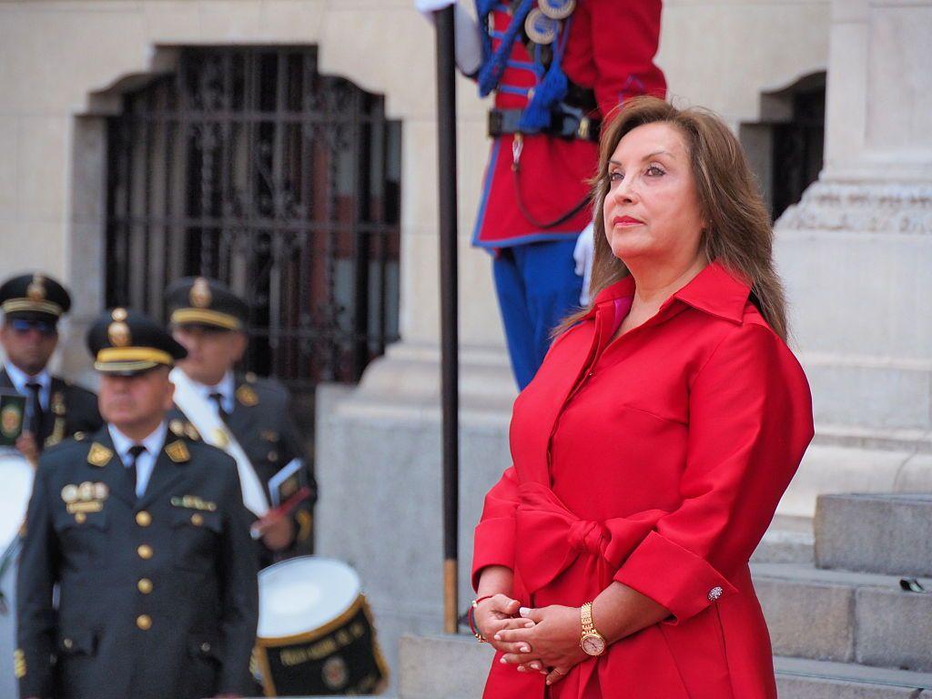 Dina Boluarte, vestida de rojo, de pie. Al fondo, un grupo de uniformados. 