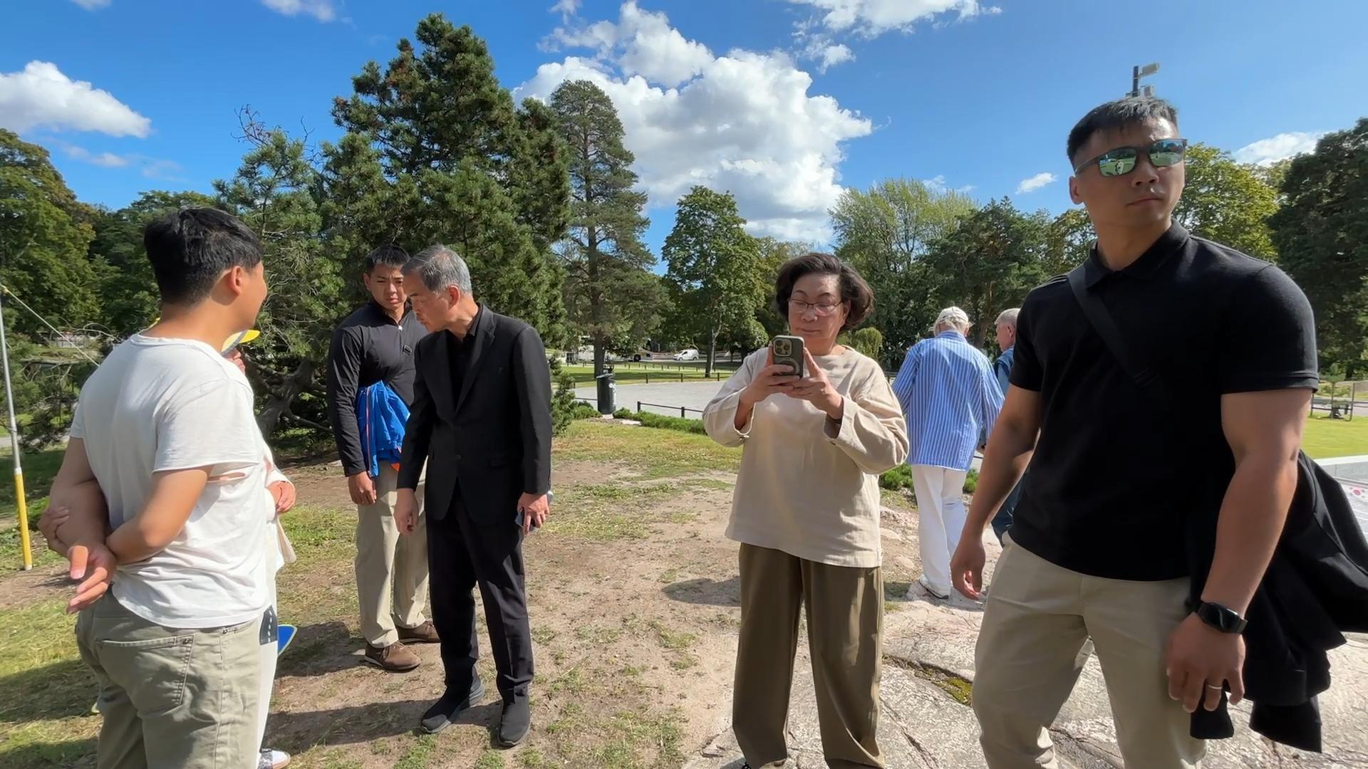 芬蘭赫爾辛基西貝柳斯公園內，身穿黑色西裝的梁振英（左四）訓斥兩名身穿白色上衣的法輪功學員（左），身穿米白色上衣的夫人梁唐青儀（右四）舉起手機疑似在拍攝拍片者，兩名身穿黑色上衣的疑似保鑣站在梁振英夫婦左右兩側（法輪大法信息中心發放影片截屏13/8/2025）