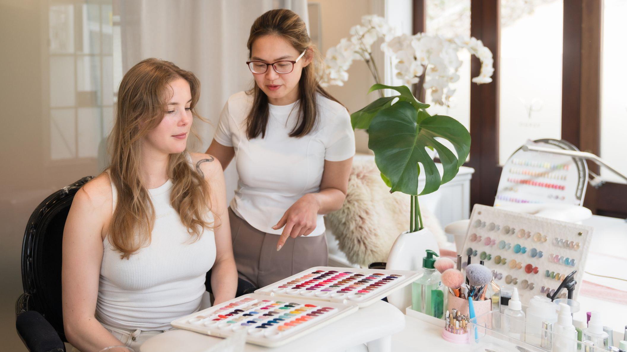 سيدة داخل أحد صالونات التجميل جالسة وهي تختار لون جل الأظافر من لوحة ألوان أظافر اصطناعية بمساعدة متخصصة أظافر خلال زيارة للصالون.