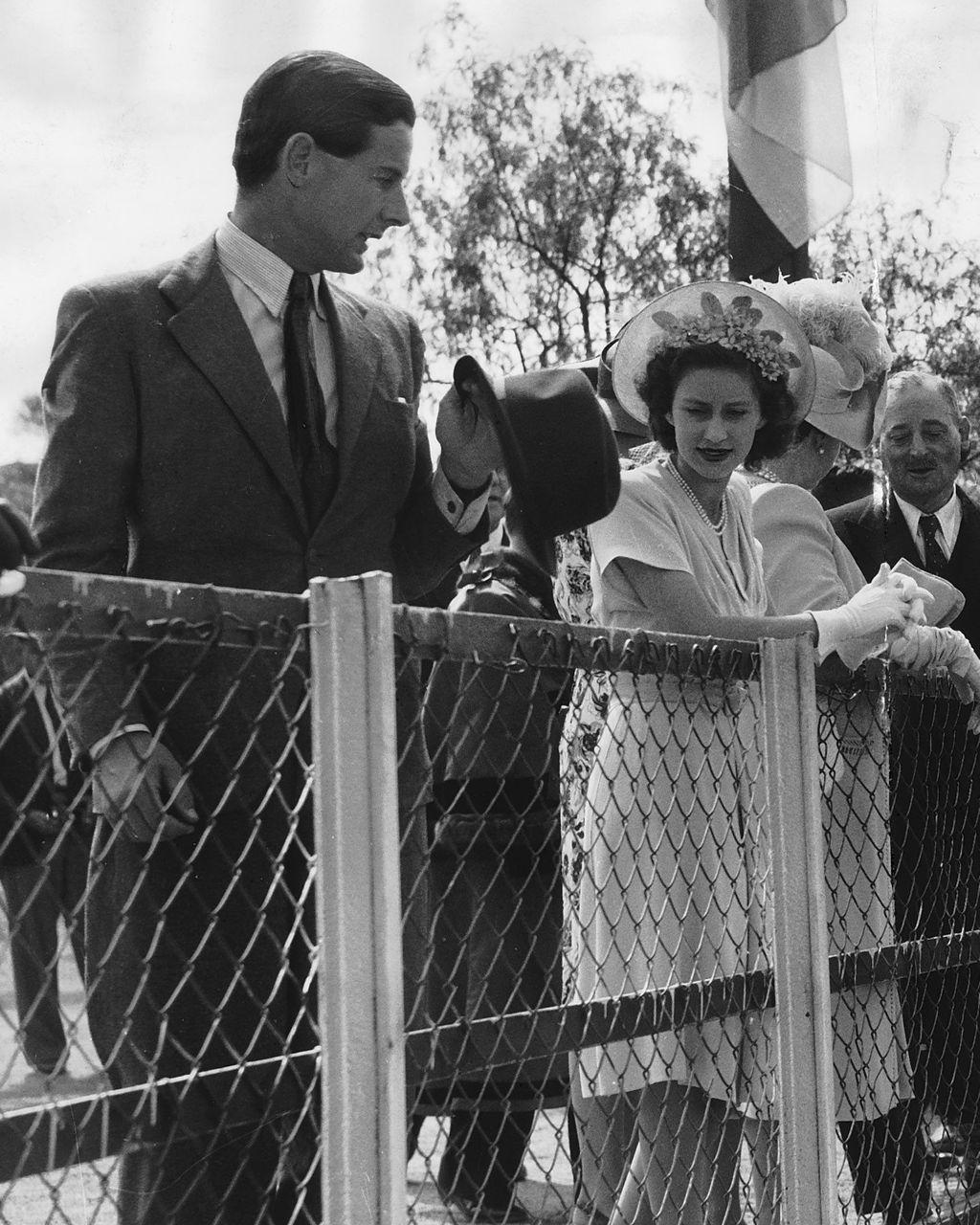 Un hombre alto con traje formal sostiene un sombrero en su mano y mira atentamente a una joven mujer vestida de blanco con un sombrero con flores. Ambos están al aire libre detrás de una verja 
