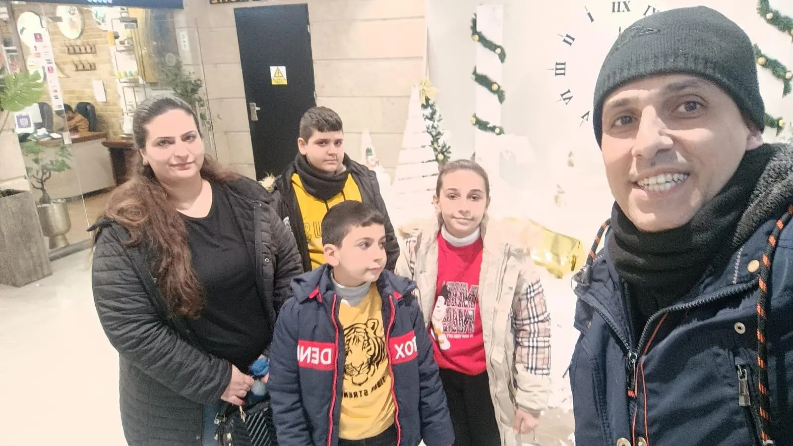 صورة لشخص من غزة يُدعى رامز الصوري برفقة زوجته وأطفاله الثلاثة مرتدين ملابس شتوية بعدة ألوان منها الأسود والأحمر والأصفر، ويظهر خلفهم واجهة زجاجية لمحل تجاري.