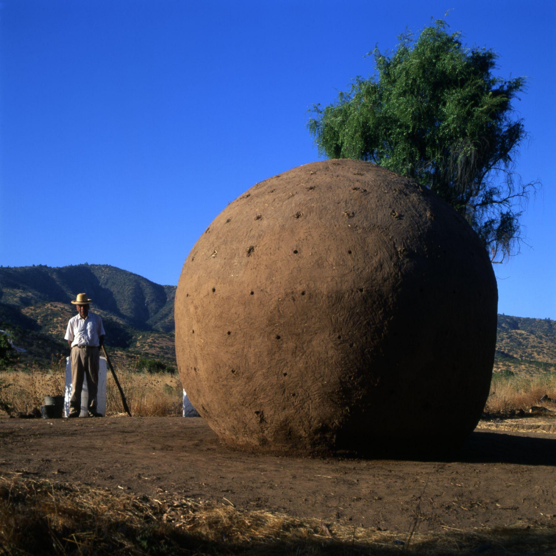 Persona de pie junto a una gran estructura esférica de tierra en un paisaje rural, con montañas al fondo y un cielo despejado.