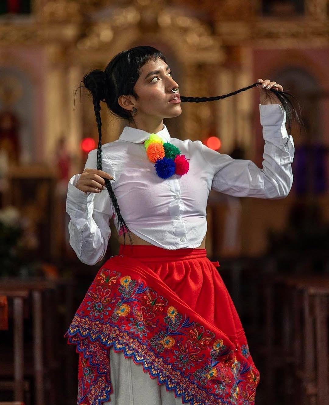 Renata Flores con una falda tradicional peruana, de color rojo, pantalones debajo, lleva una camisa blanca con pompones de colores. 