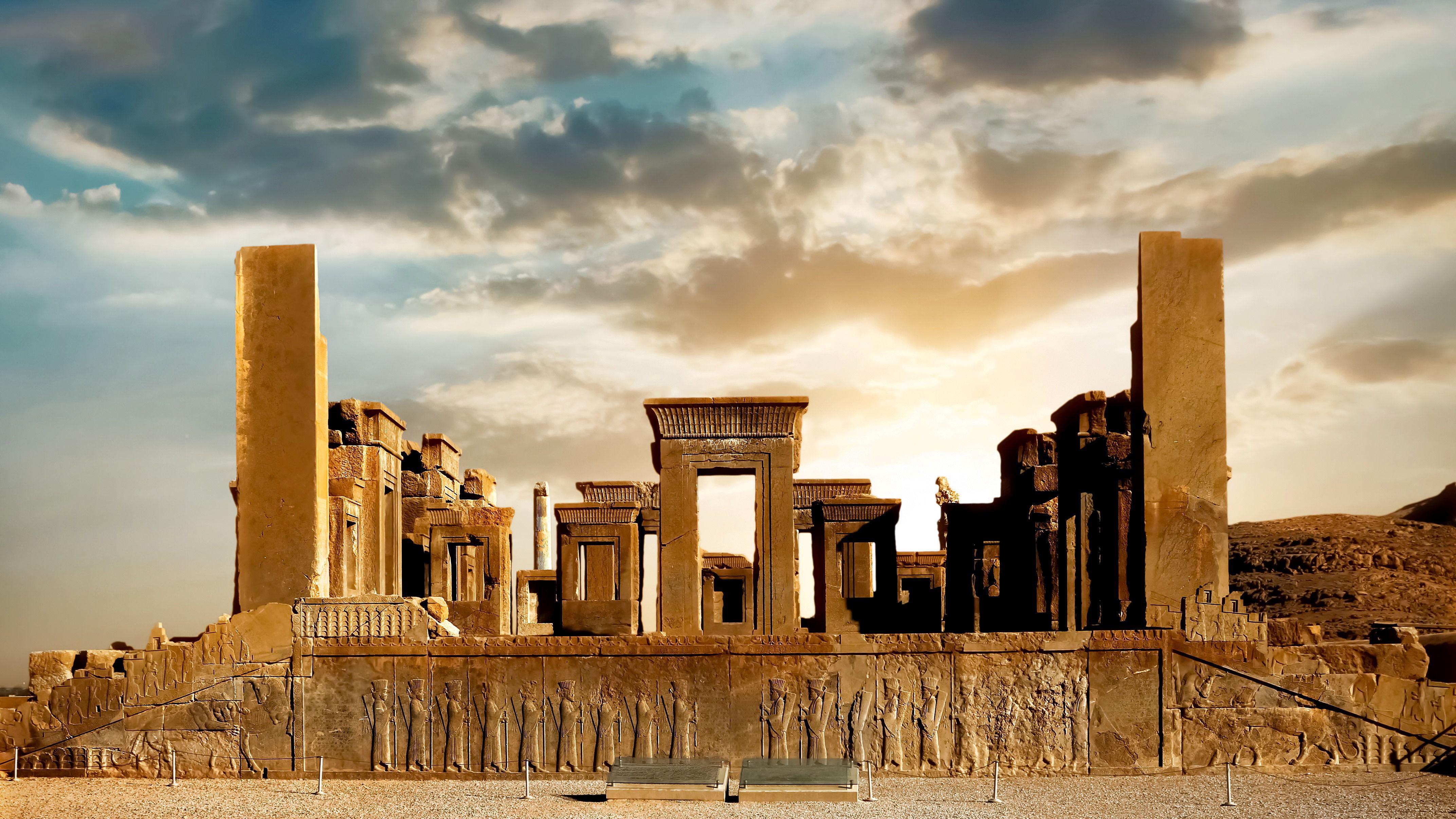 El sol sale sobre las escaleras y columnas de piedra tallada de la terraza de la antigua ciudad persa en ruinas de Persépolis. Una zona elevada de piedra alberga numerosas columnas gigantes y estructuras adinteladas. Hay figuras talladas en la base de la plataforma.