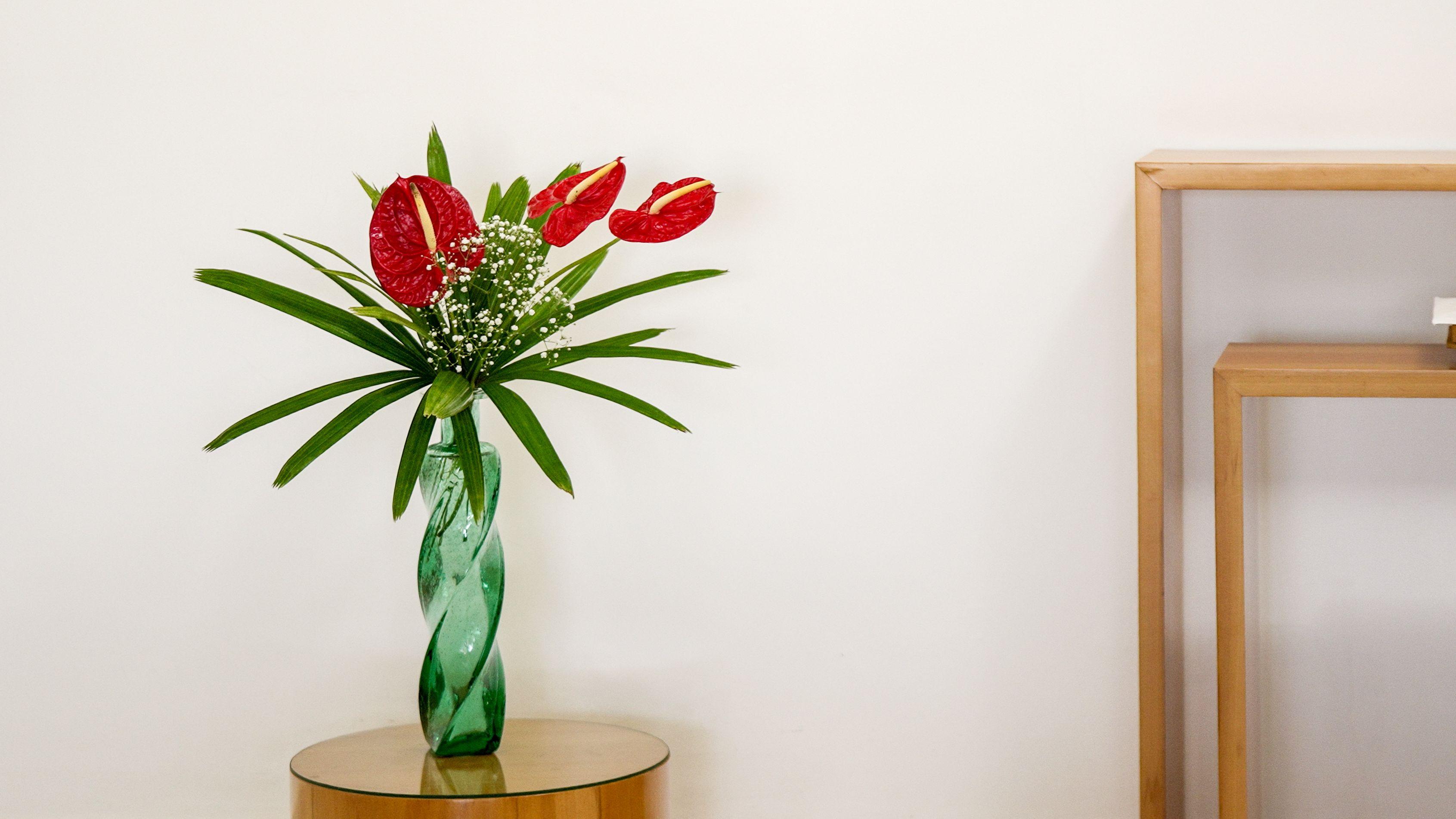 Imagem de um vaso fino verde de vidro com um arranjo minimalista de flores vermelhas e uma folhagem sobre uma mesa redonda pequena. 