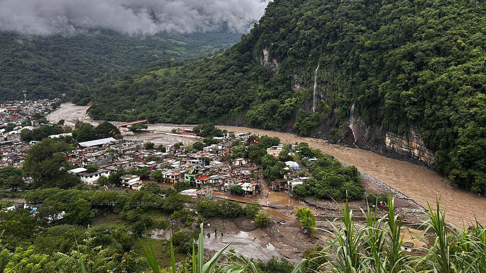 Vista general de una aldea al pie de una montaña, con casas de diferentes colores, está inundada por un río desbordado. En el fondo se ven nubes grises