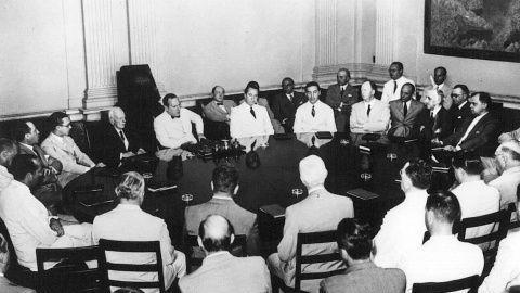 Fotografia em preto e branco de uma reuni&atilde;o da Cepal (Comiss&atilde;o Econ&ocirc;mica para a Am&eacute;rica Latina). V&aacute;rios homens, a maioria de terno e gravata, est&atilde;o sentados ao redor de uma grande mesa oval, com um quadro ao fundo.