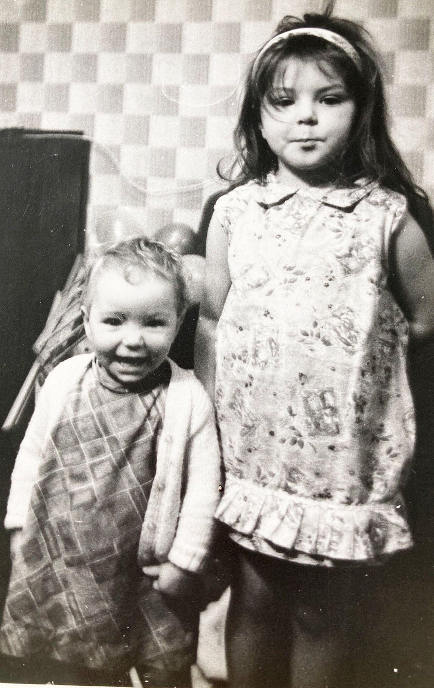 Uma fotografia em preto e branco de duas meninas dentro de casa. A mais velha est&aacute; em p&eacute; usando um vestido estampado com uma tiara, enquanto a mais nova est&aacute; ao lado, sorrindo, vestindo um conjunto xadrez e um cardig&atilde;. Atr&aacute;s delas h&aacute; uma parede com um papel de parede de padr&atilde;o quadrado distinto e alguns bal&otilde;es
